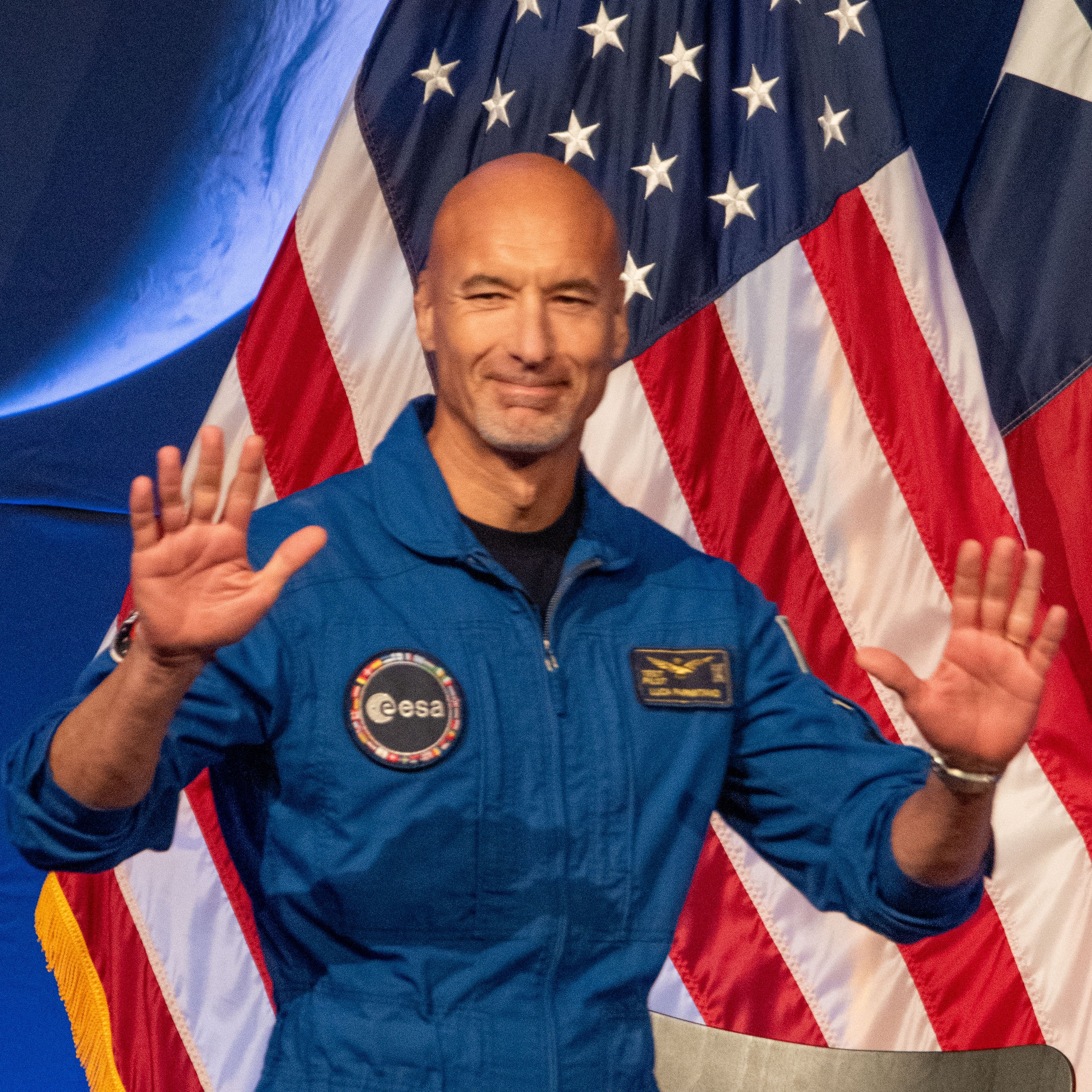 Um homem careca de macacão azul levanta as mãos.