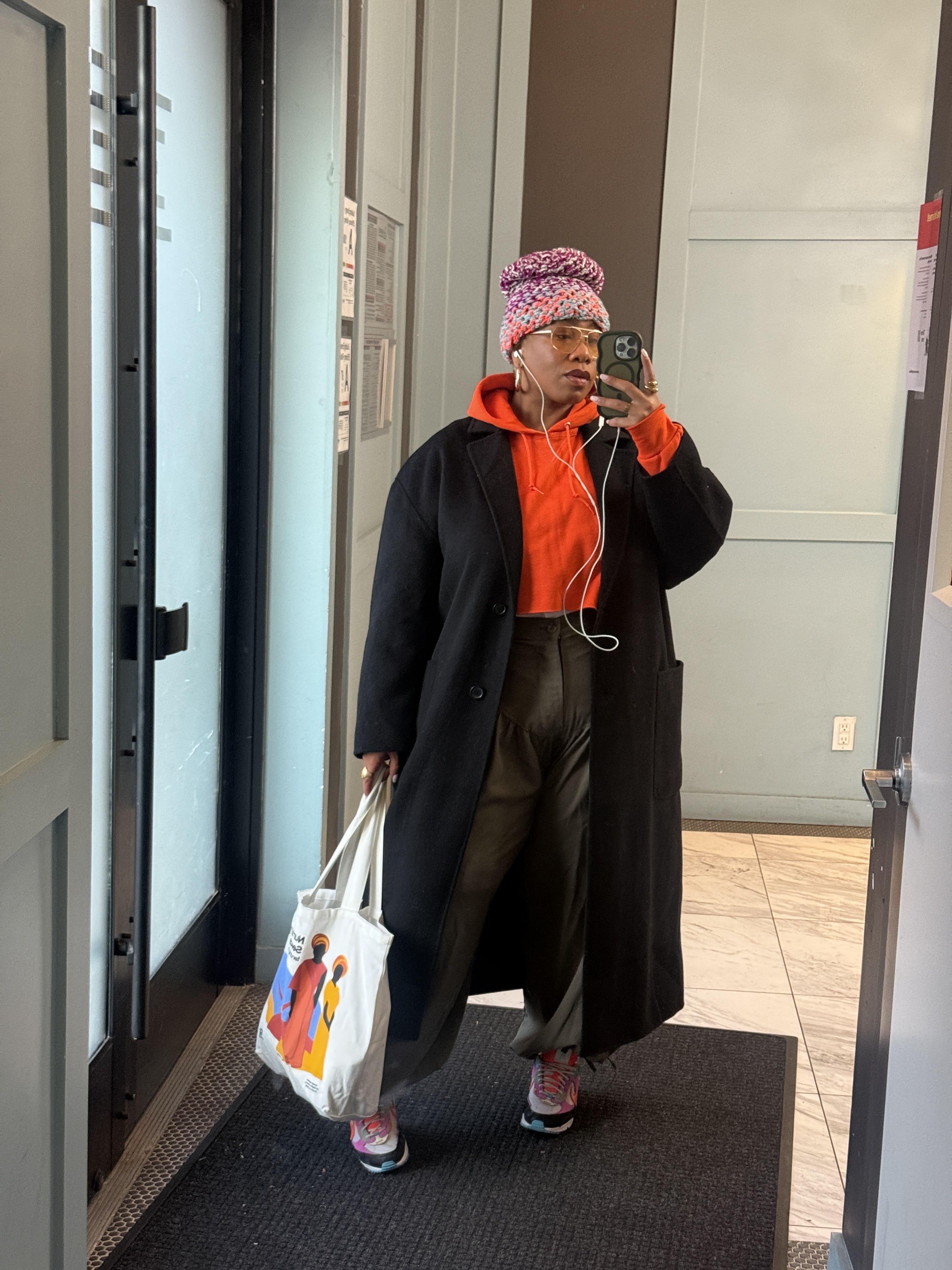 Kelly Augustine wearing a black coat, a red hoodie, and a print hat with a tote bag