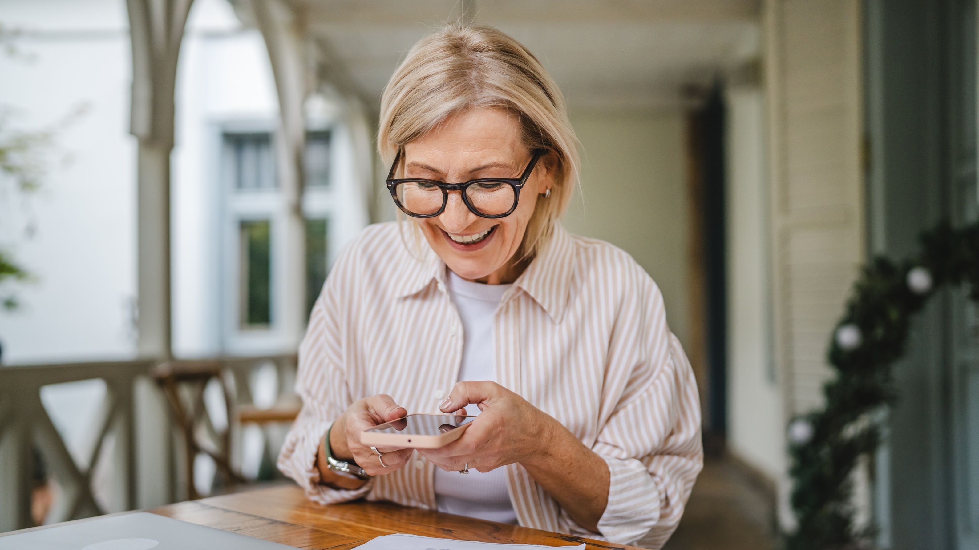 An older woman smiles as she looks at her phone while sitting at a table on her porch.