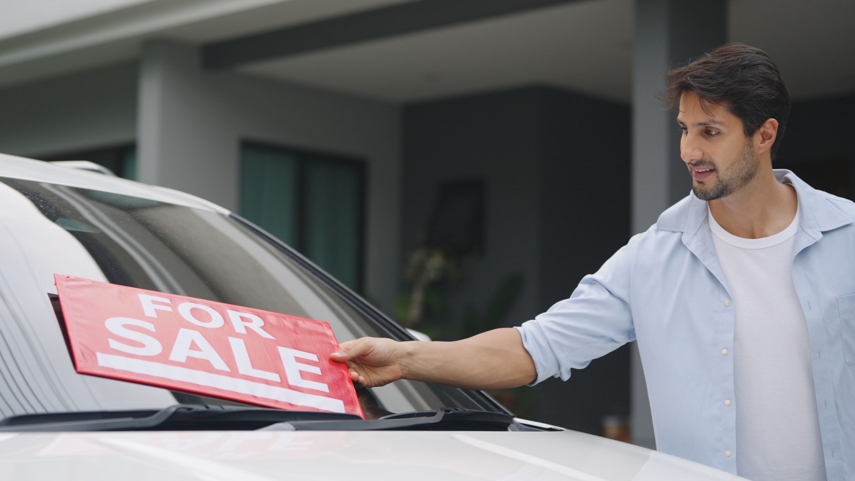 A young man placing a red "For Sale" sign on the windshield in front of a car