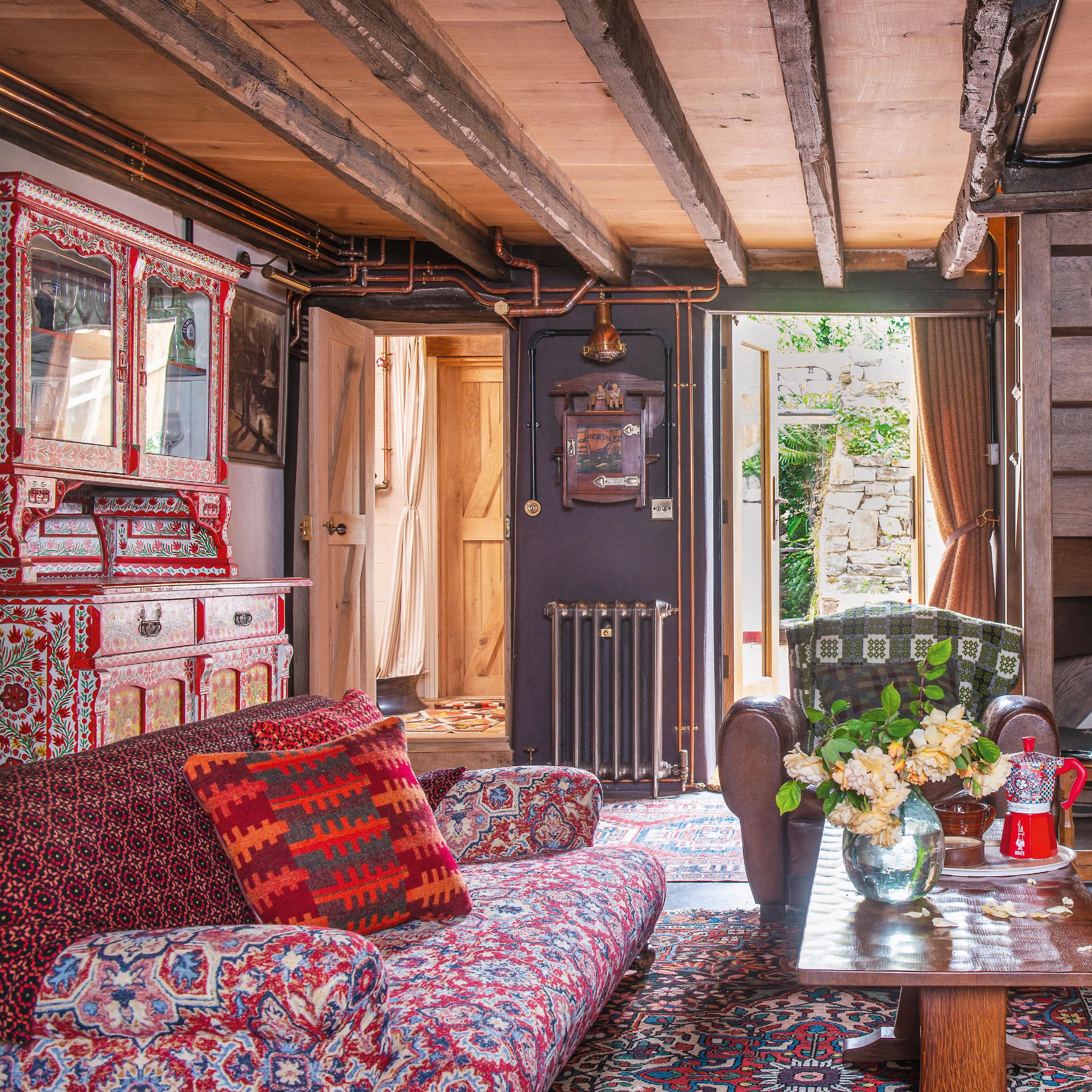 living room in rustic cottage with exposed ceiling beams and wall panelling with patterned sofas, blankets and antique furniture