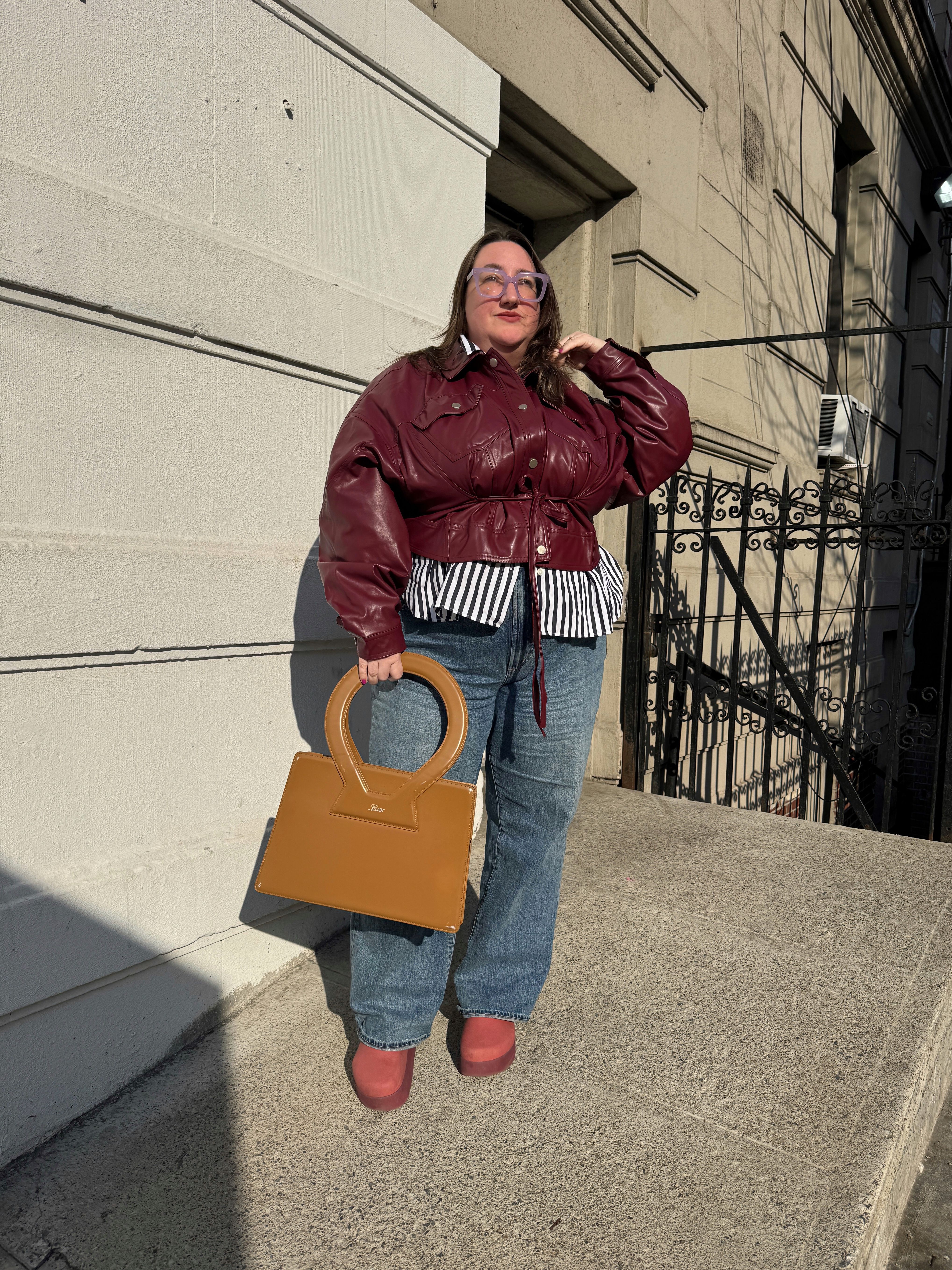 Sarah Chiwaya styles a cinched-waist merlot leather jacket over a button-down and jeans, with an oversized handbag