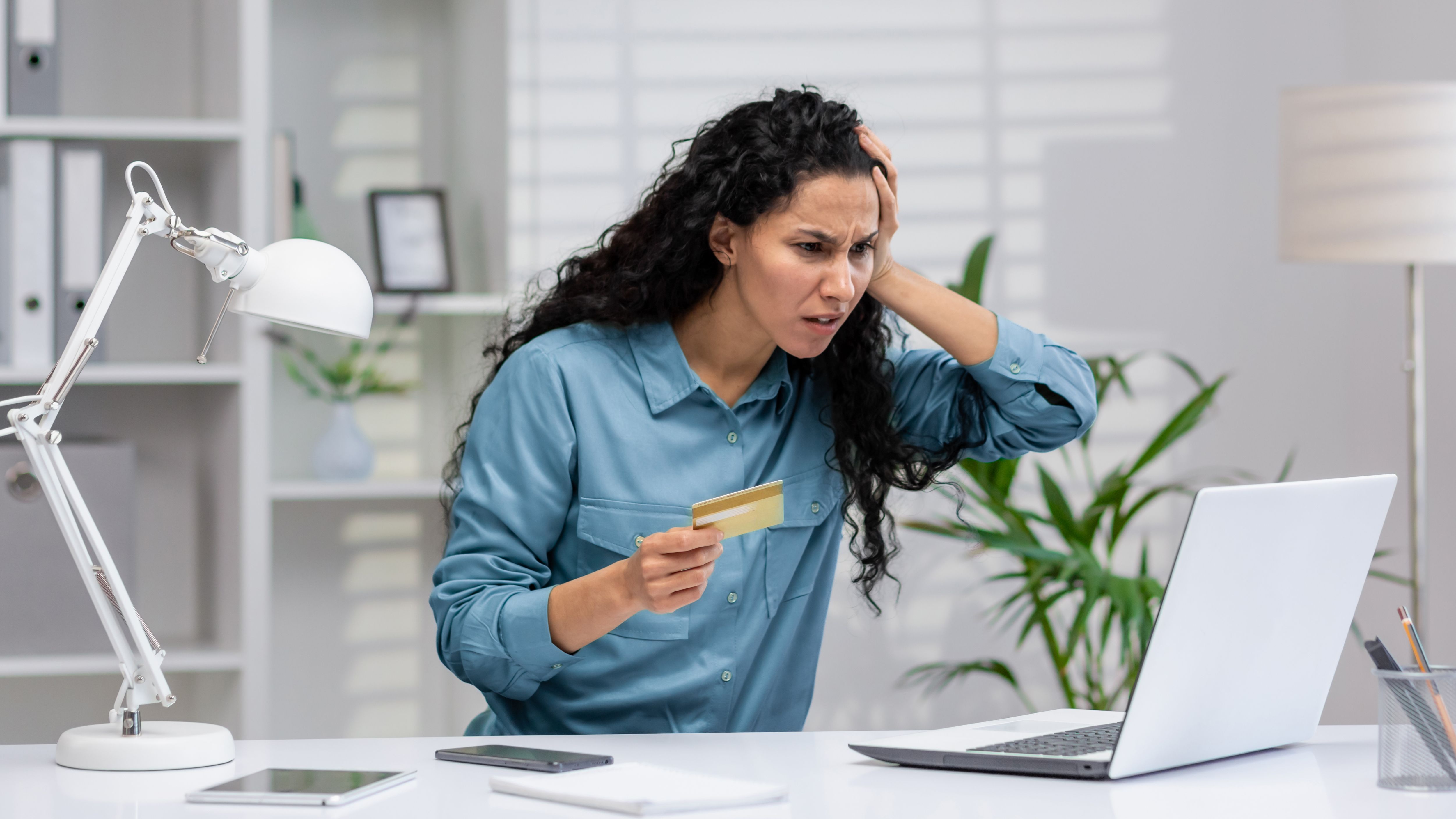Woman sat at an office desk in front of a laptop holding a credit card looking stressed