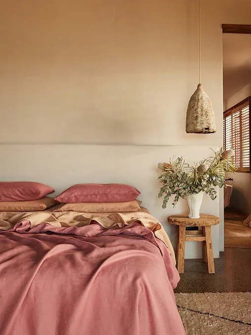 A warm, earthy bedroom featuring blush-pink linen bedding, natural wood accents, a rustic stool used as a bedside table, a textured pendant light, and soft draped curtains filtering natural light.