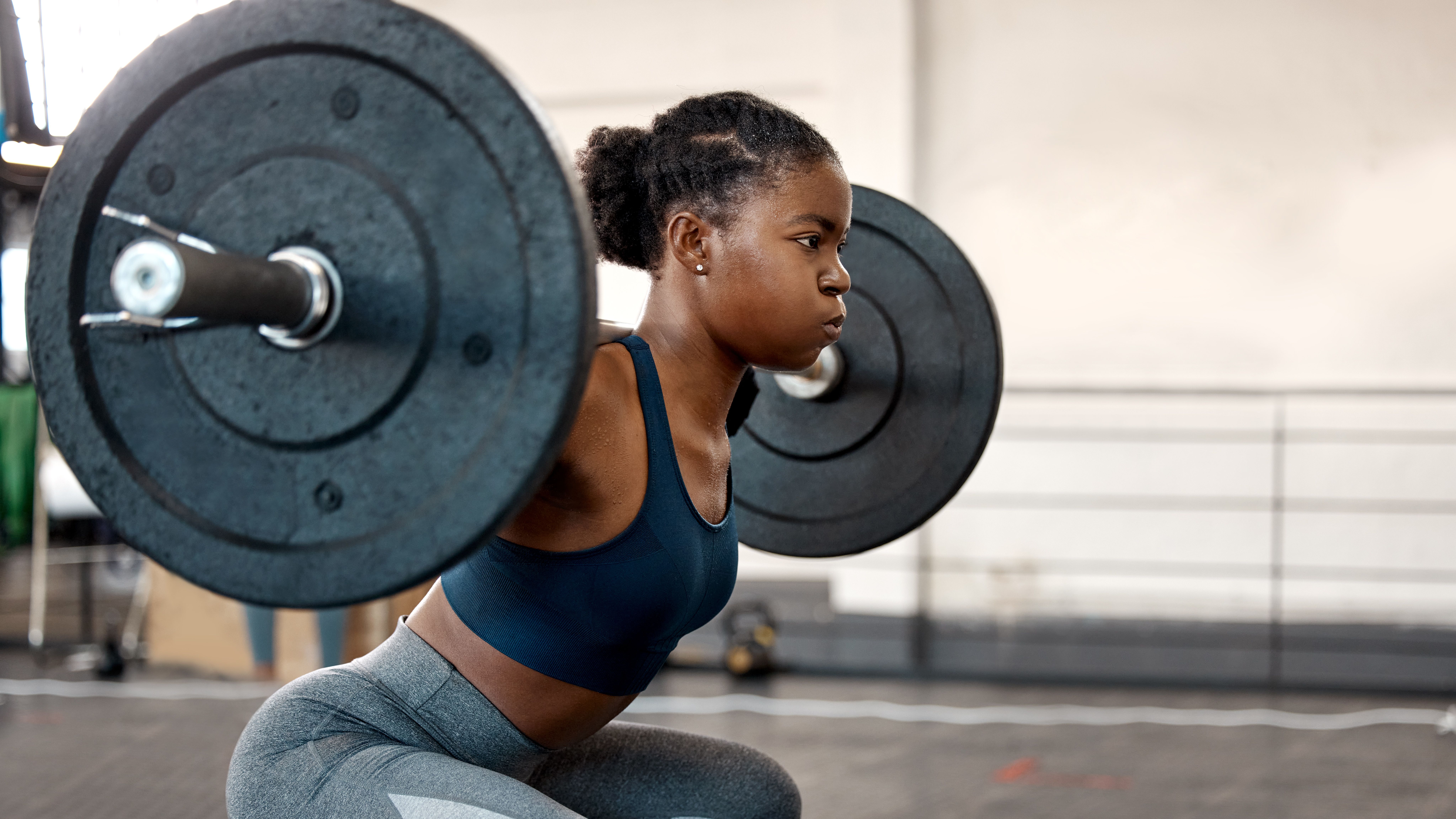 A woman performs a barbell squat, wearing a sports bra and leggings. She is squatting down, with her knees bent, and a barbell positioned across her back.