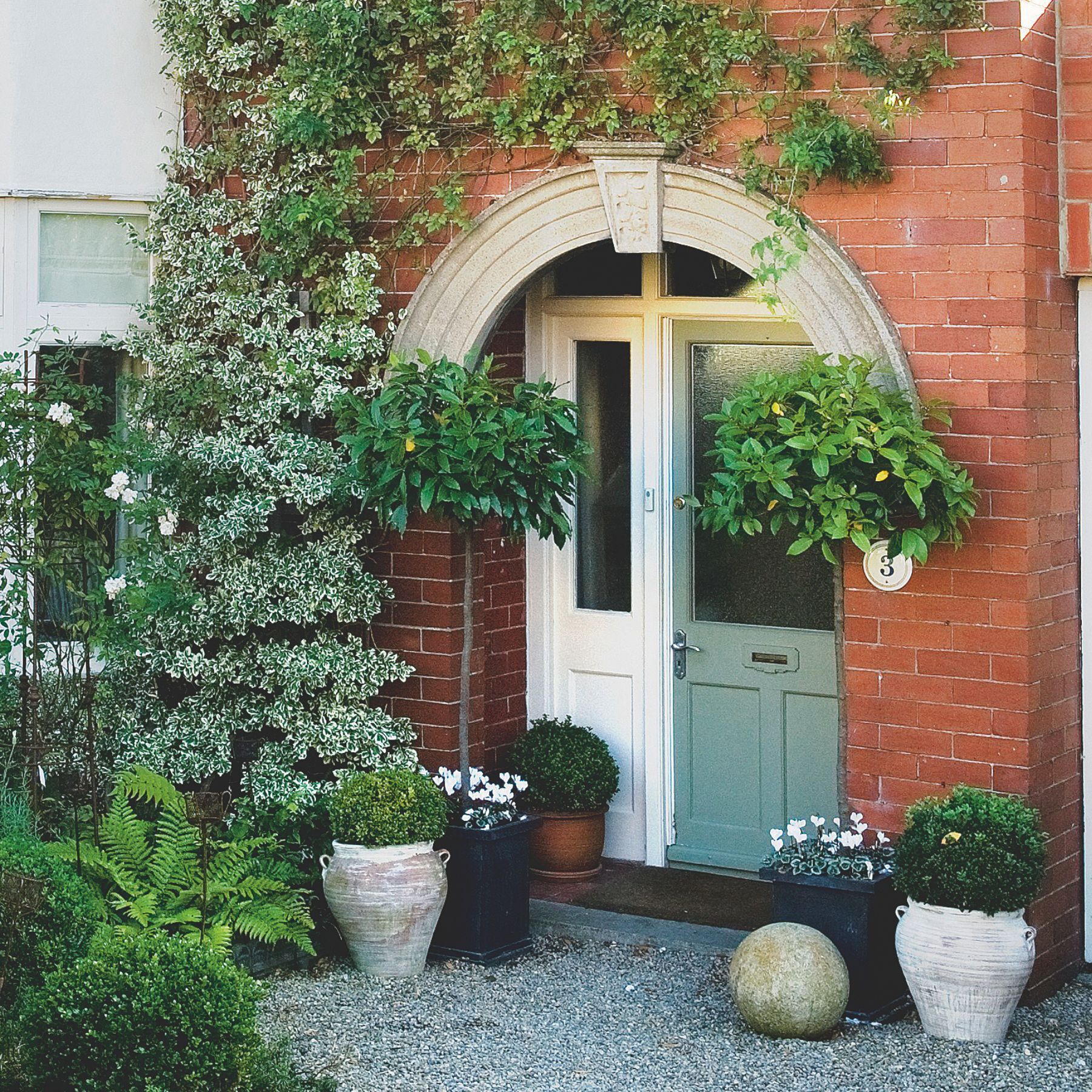 Exterior of brick house with matching colour front door and garage door surrounded by established plants and pretty pots