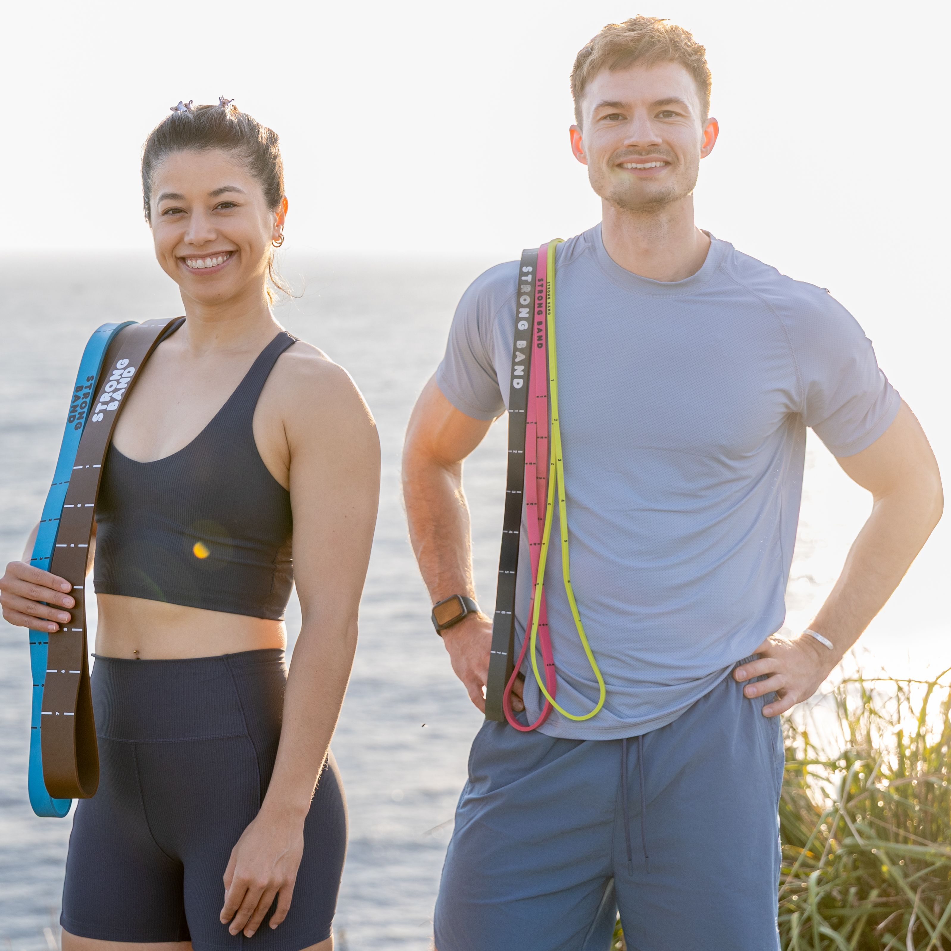 Man and woman stand outside with sea behind them. They have multiple resistance bands draped over one shoulder