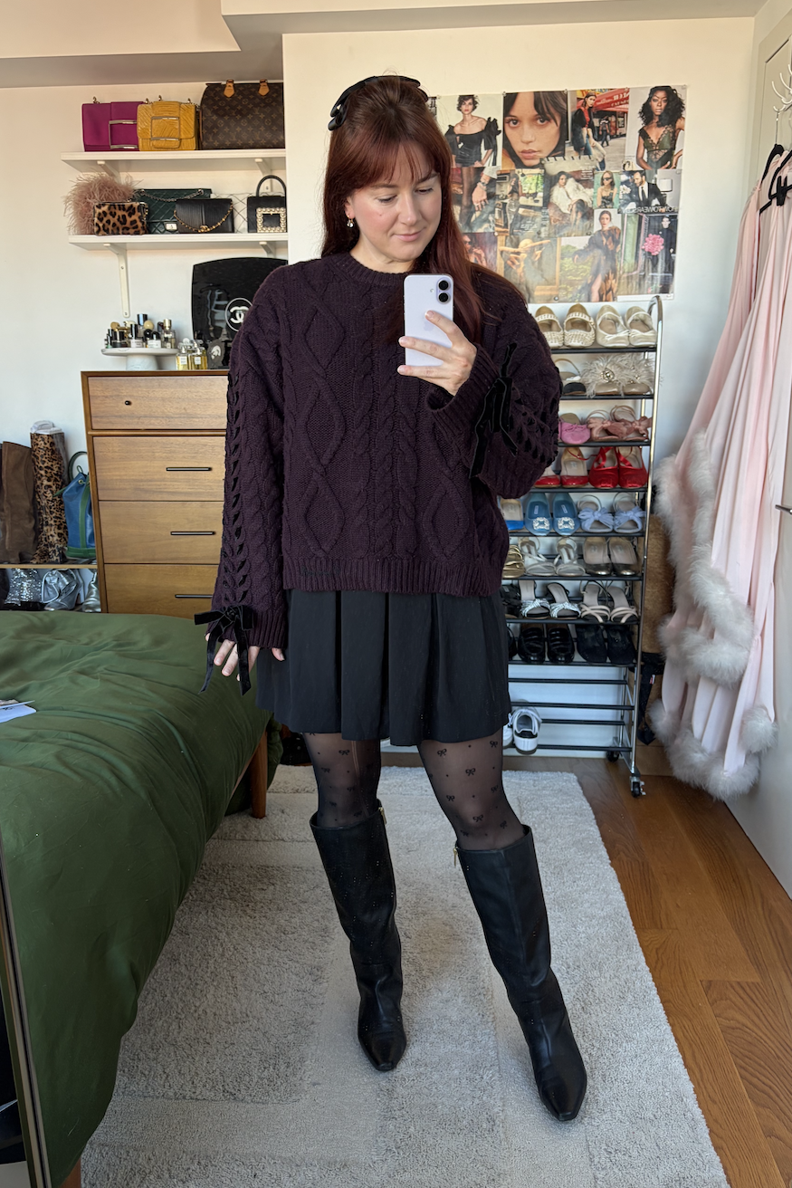 Tyler McCall wearing a purple cable-knit sweater, black pleated skirt, polka dot tights, and black leather knee-high boots