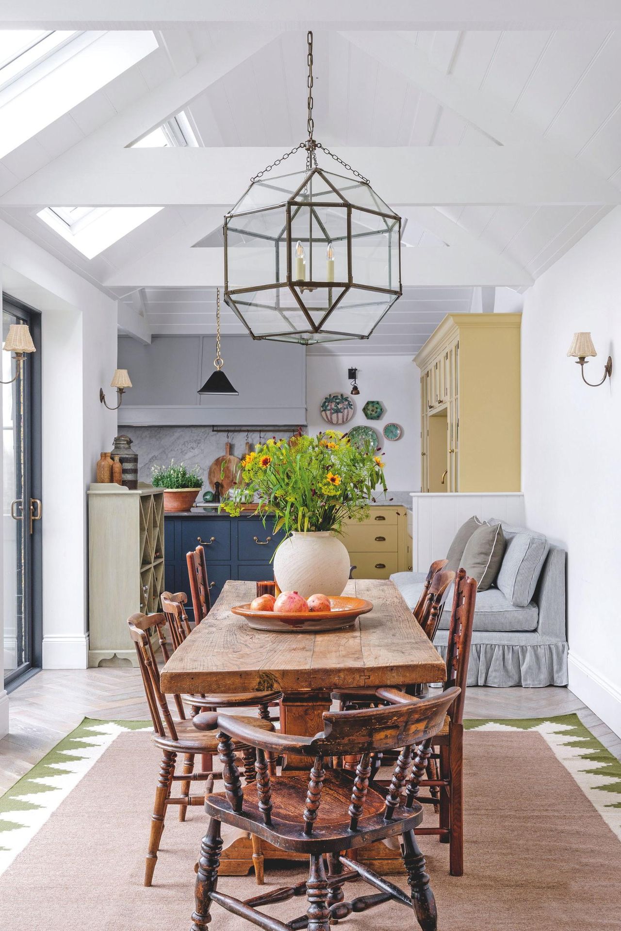 A bright, airy dining space with a rustic wooden table, antique spindle chairs, and a large glass lantern pendant light hanging from a vaulted white ceiling.