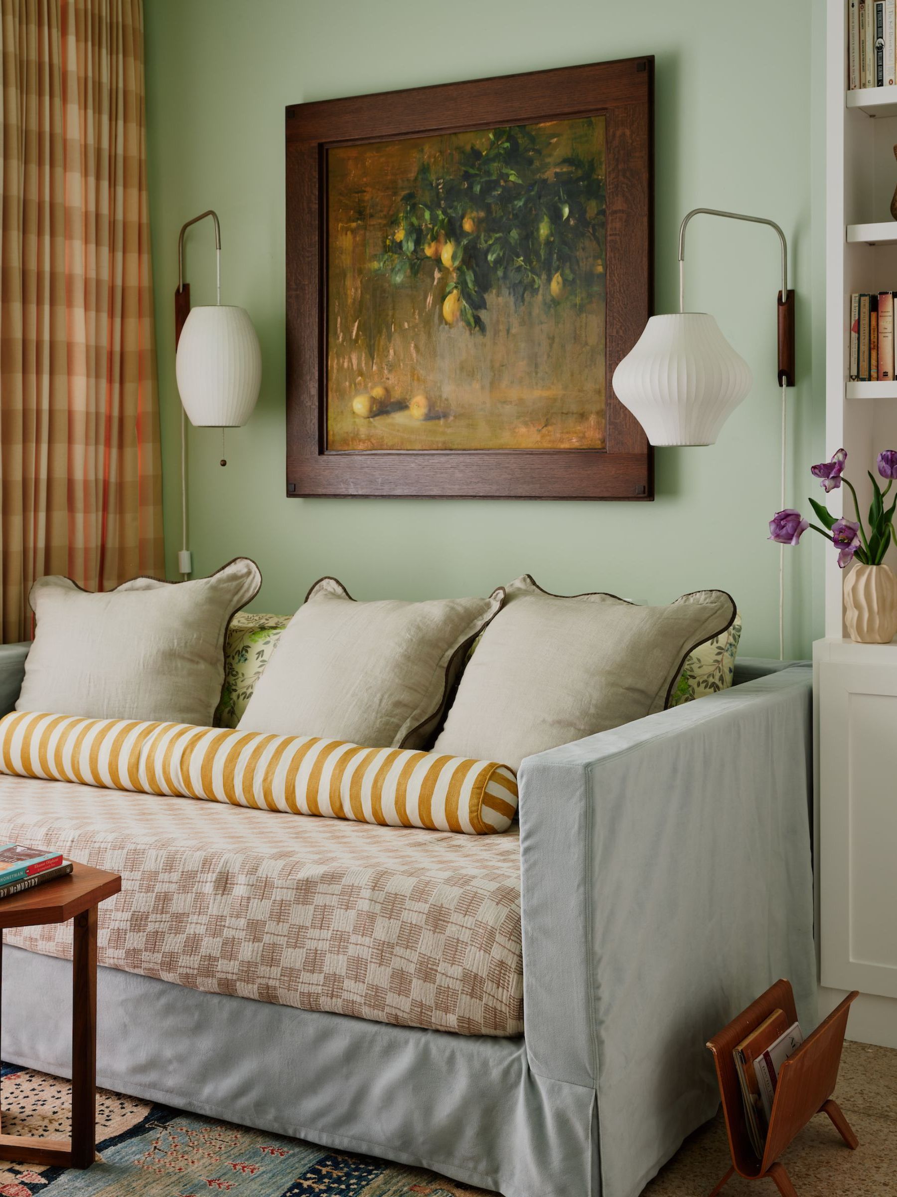 A guest home office with pale green walls, a sofa bed with layered textiles in soft green, ochre, and pink. Checked orange and pink curtains, vintage artwork on the wall, and two modern wall lamps.