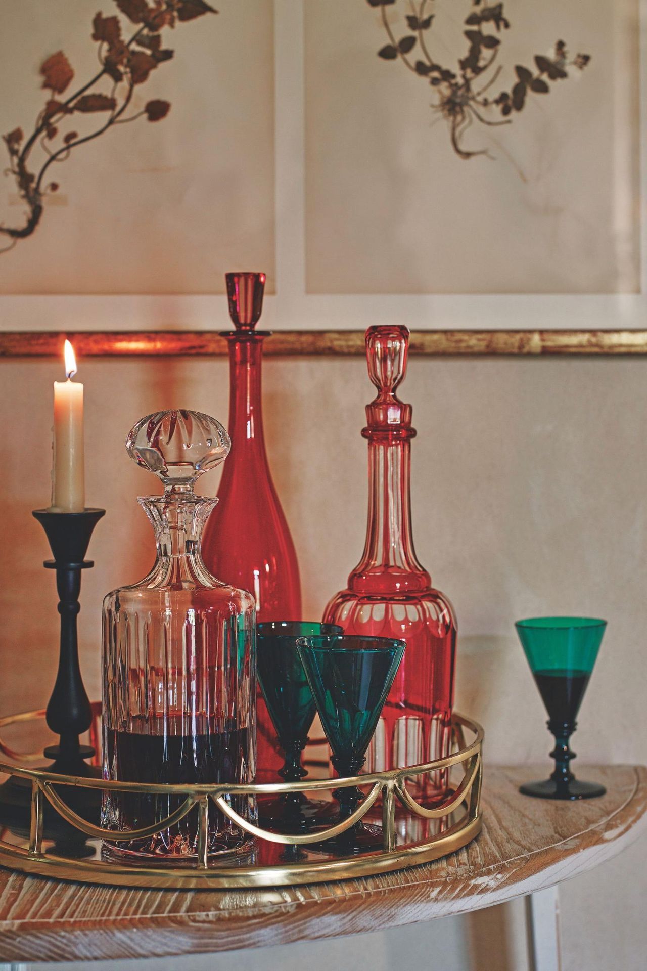 Vintage colored glassware on a gold mirrored tray on a wooden table. There is a lit taper candle, and above are two vintage-look botanical prints in a gold frame.