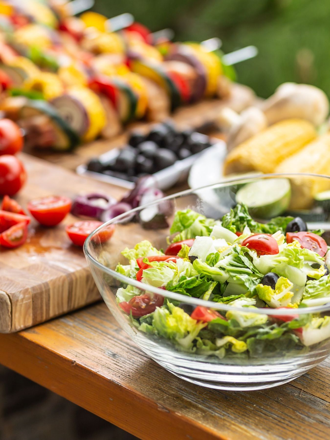 Garden table full of food ready for a barbeque party with a salad in the front and a woman cutting vegetable aside.