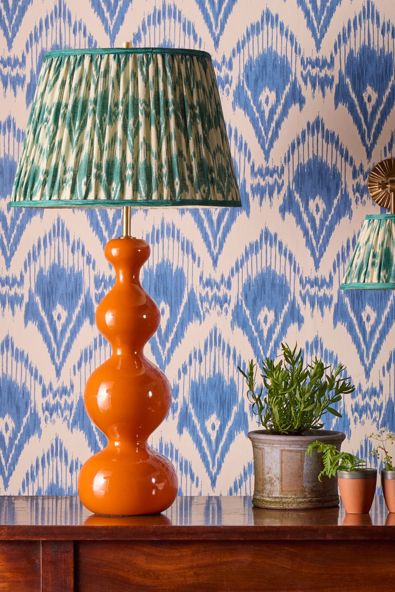 Sculptural orange GP&amp;amp;J Baker x Pooky Lighting table lamp with a blue patterned lamp shade pictured in a room with ikat wallpaper and various potted plants.