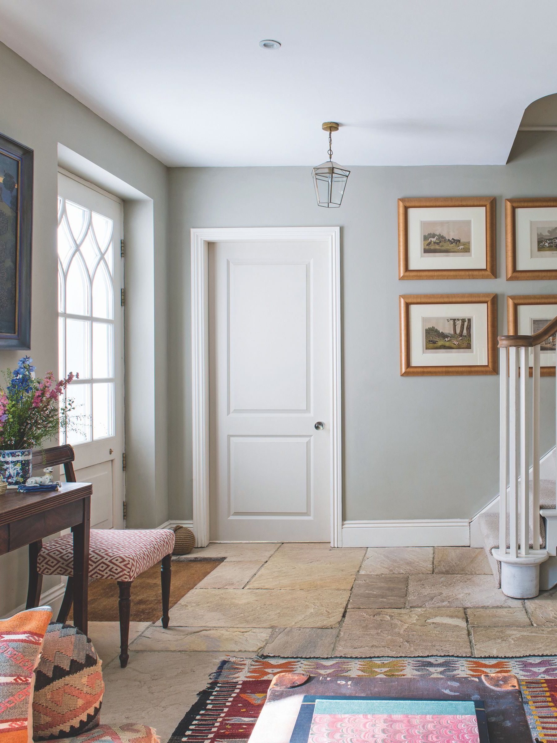 A bright and colorful entryway with tiled flooring, grey walls, patterned rug and cushions, wooden console table with flowers and framed wall art with white staircase and white painted door