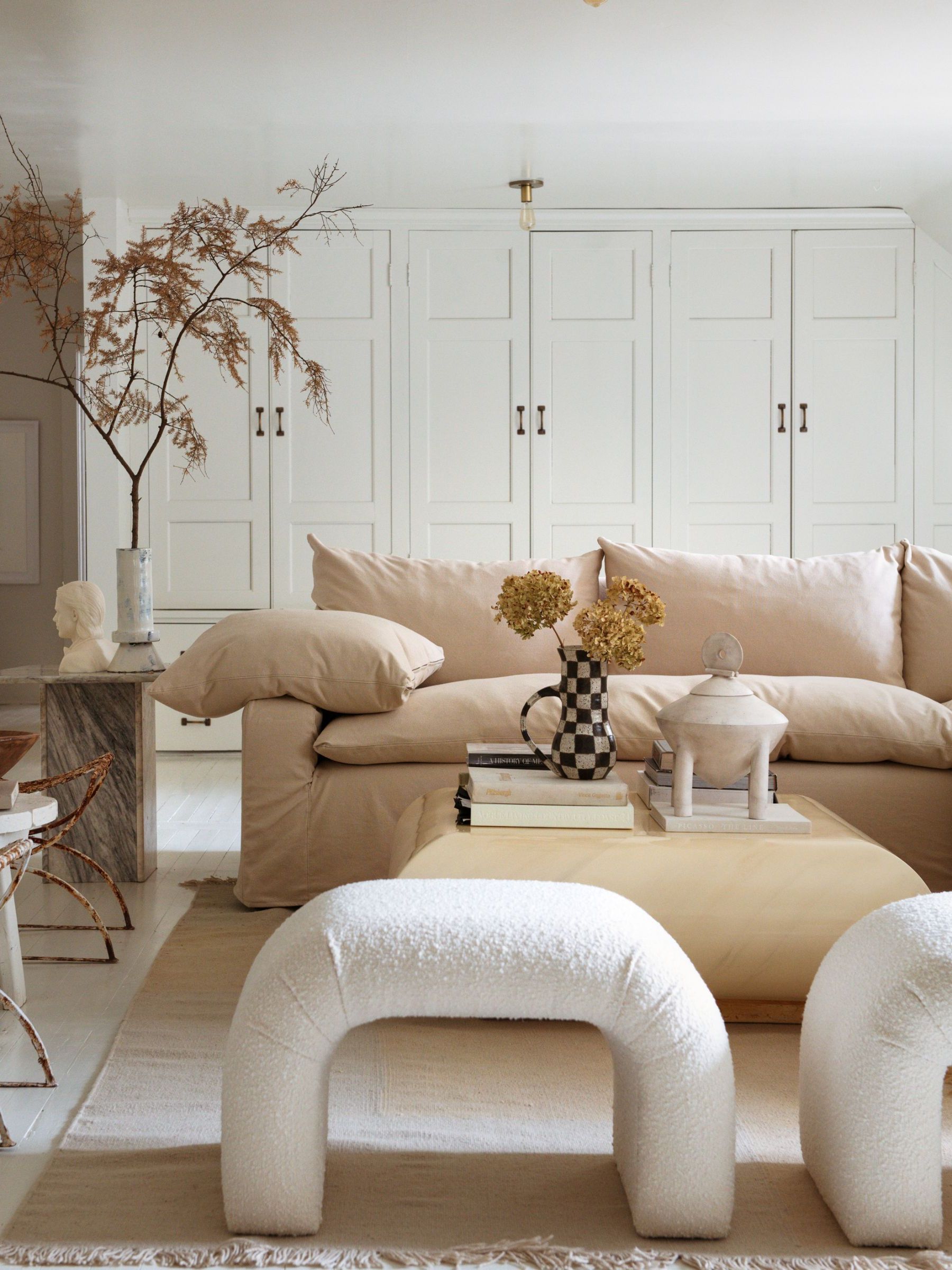 A light and airy living room with white walls and built-ins, beige sofa, pale wood coffee table, a neutral rug and two modern boucle seats.