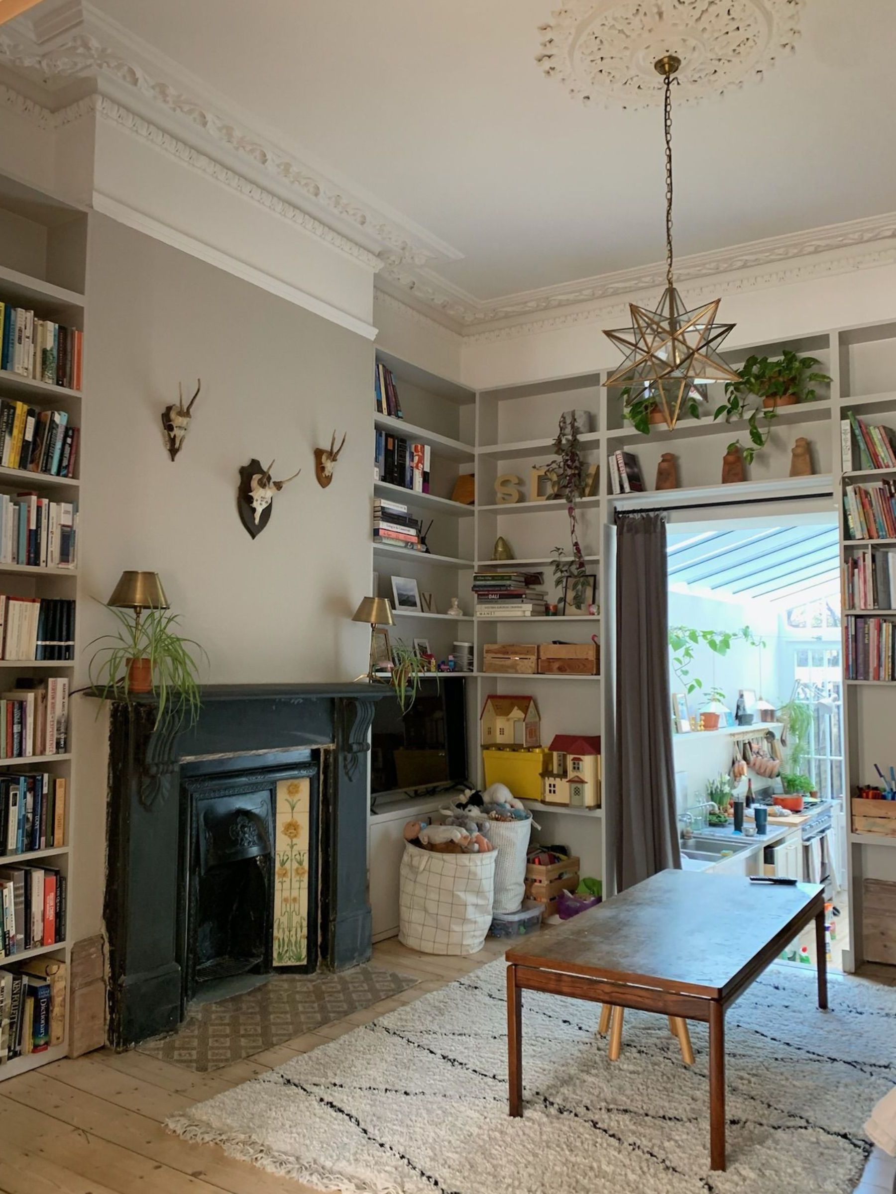 Living room with built-in shelving, grey painted walls, white ceiling with crown molding and ceiling medallion, star lantern hanging from the ceiling, Berber rug, and a large Victorian fireplace