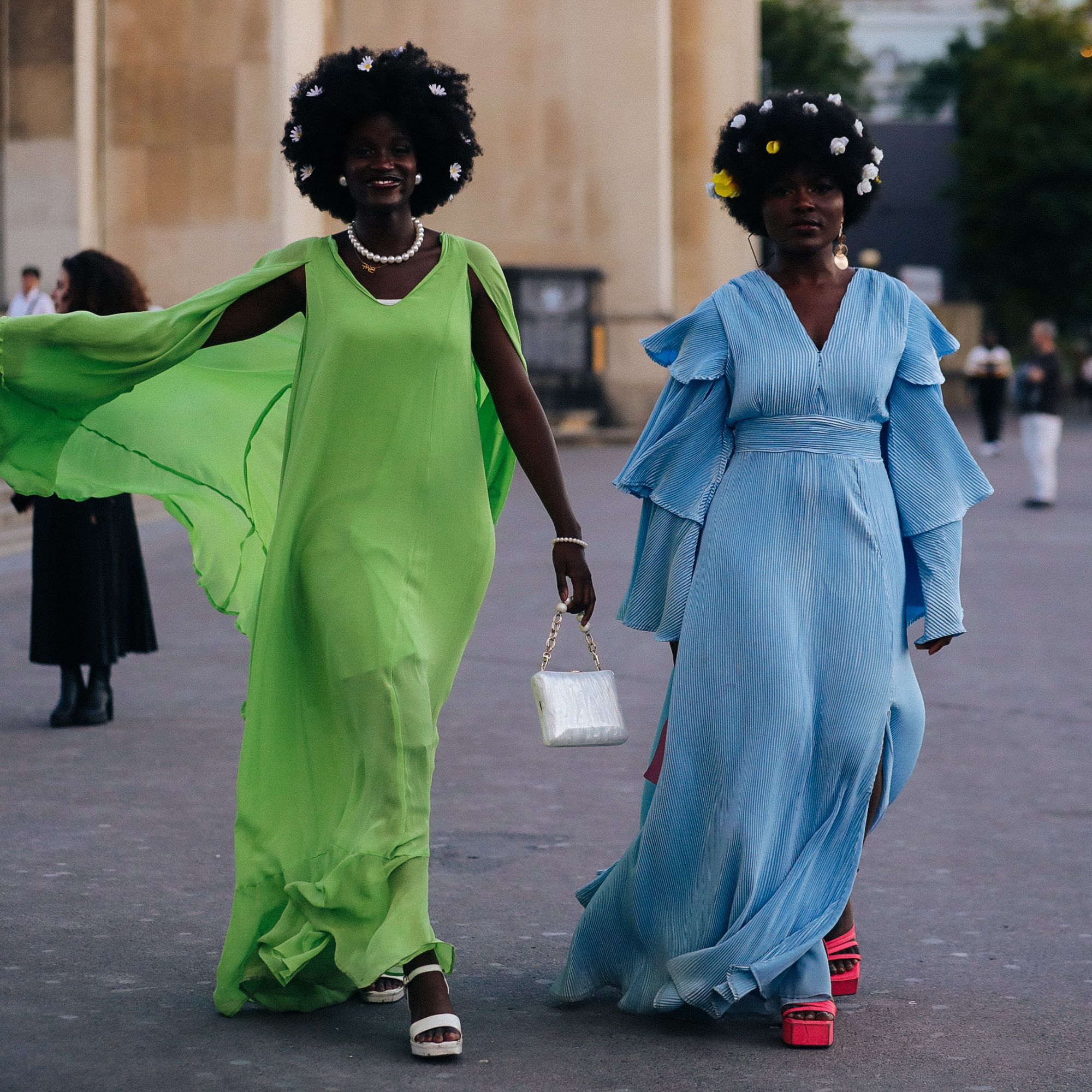 Two women with wearing small flowers in their coily hair. One woman is wearing a green dress and the other is wearing a blue dress.