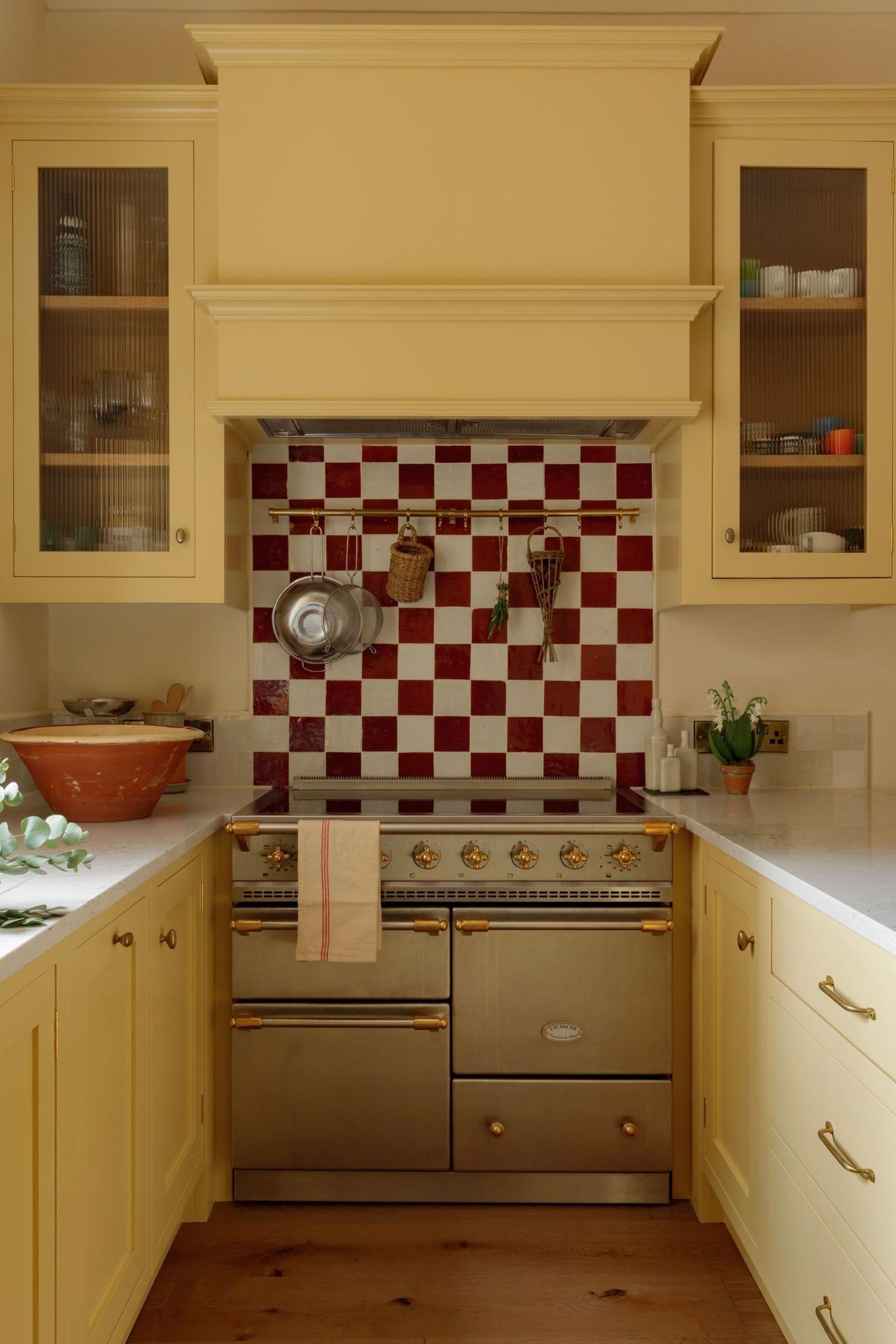 Small Georgian kitchen with Shaker cabinets in yellow, red and white checkerboard backsplash, brass range cooker, and tall glazed storage units.