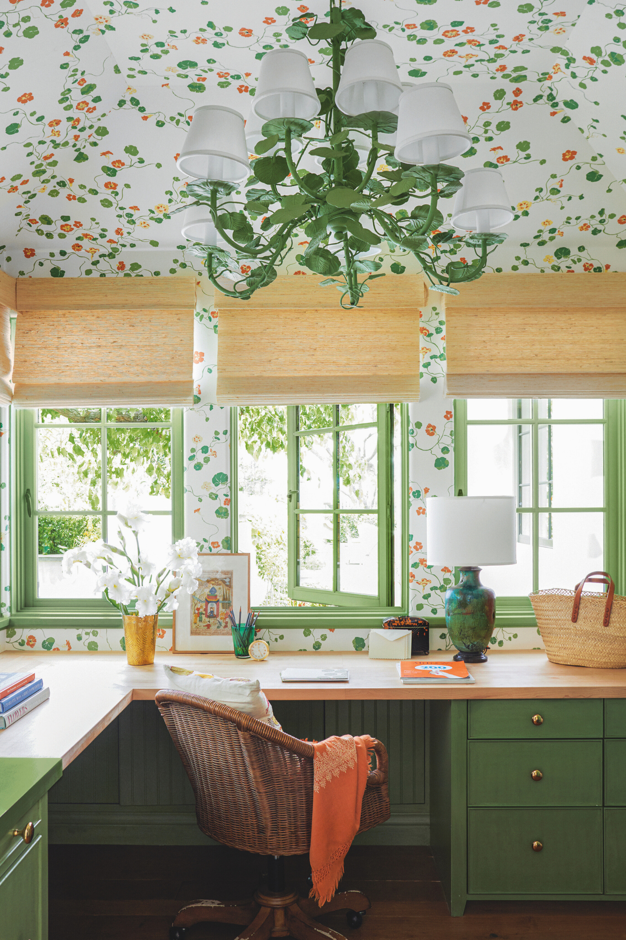 a home office with a built in countertop with green painted drawers, decor on the countertop surface, windows with blinds in front of them, and wallpaper in a floral pattern that continues up to the ceiling