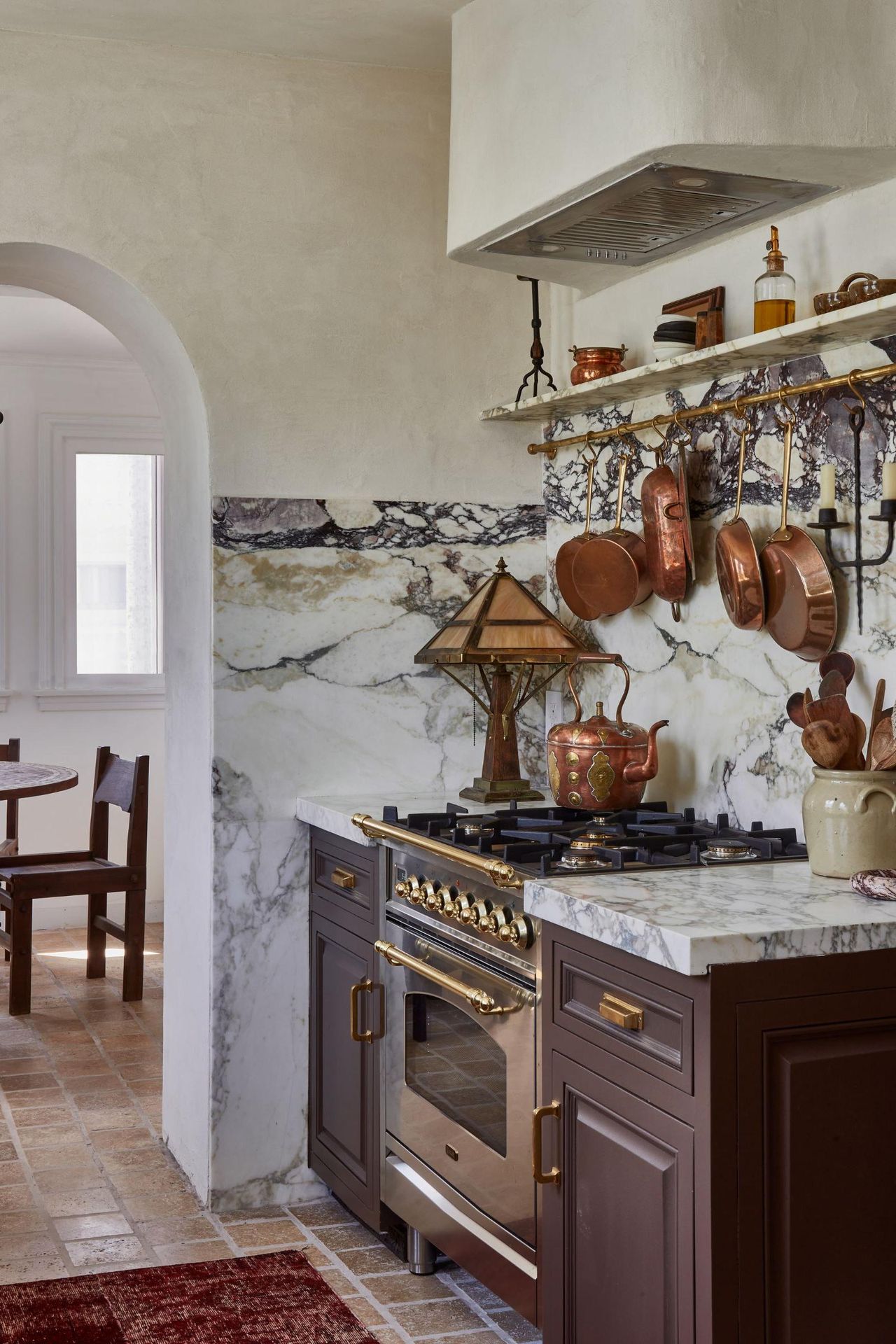A Vintage kitchen with brown cabinets, a marble countersplash, a brass pot rack on the wall, and a range cooker topped with a vintage copper teapot