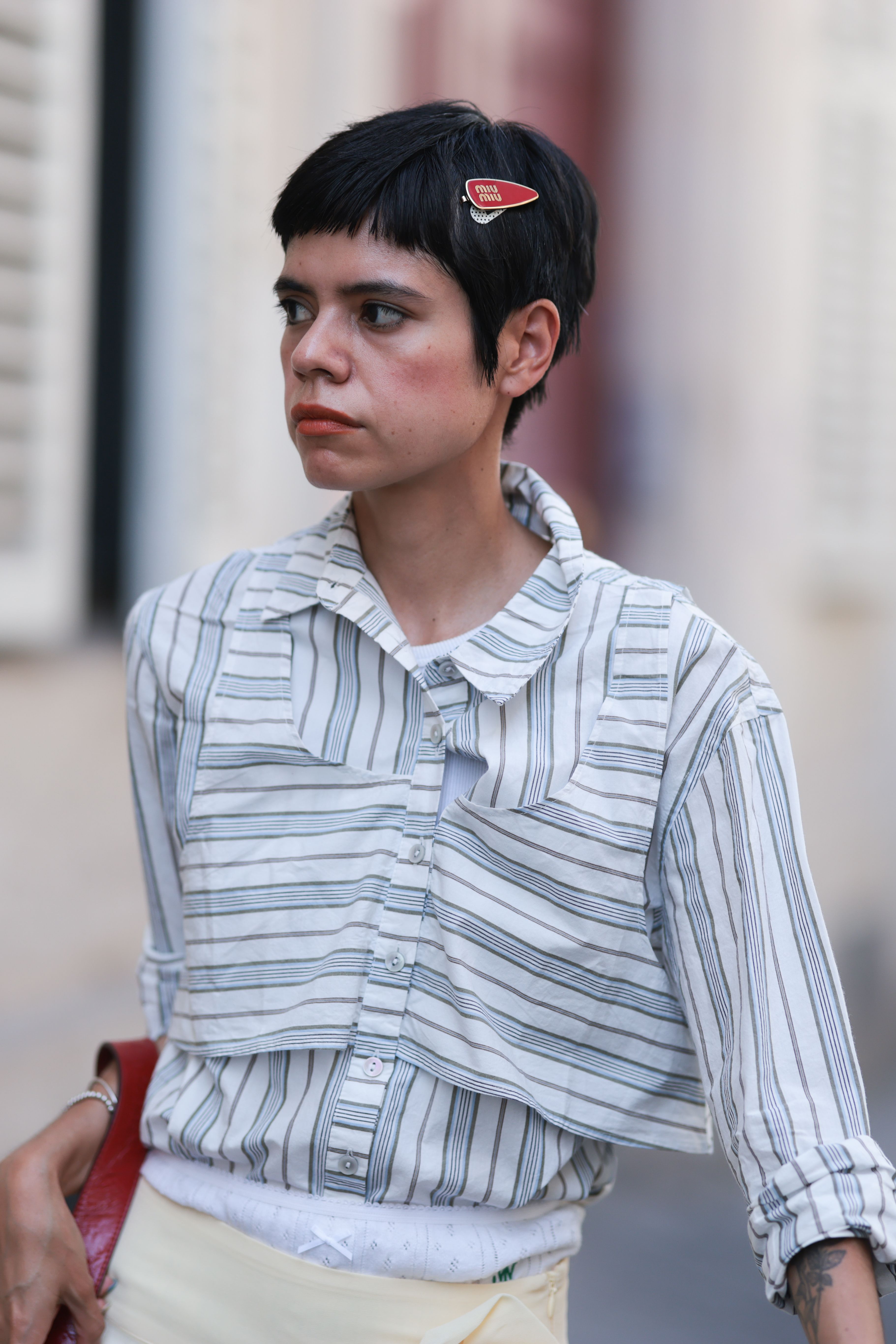 A woman wearing a red beret in her pixie hairstyle. She is also wearing a striped button up shirt and is holding a red bag.
