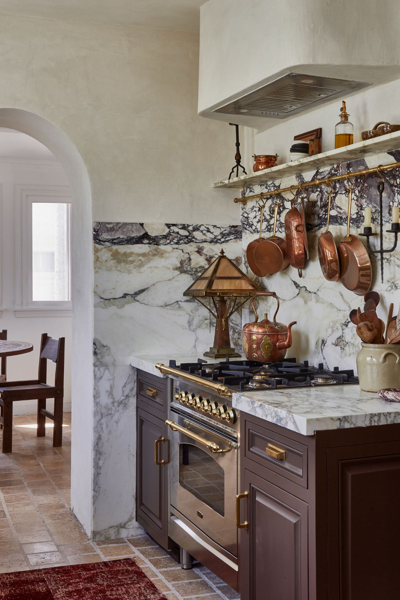A Vintage kitchen with brown cabinets, a marble countersplash, a brass pot rack on the wall, and a range cooker topped with a vintage copper teapot