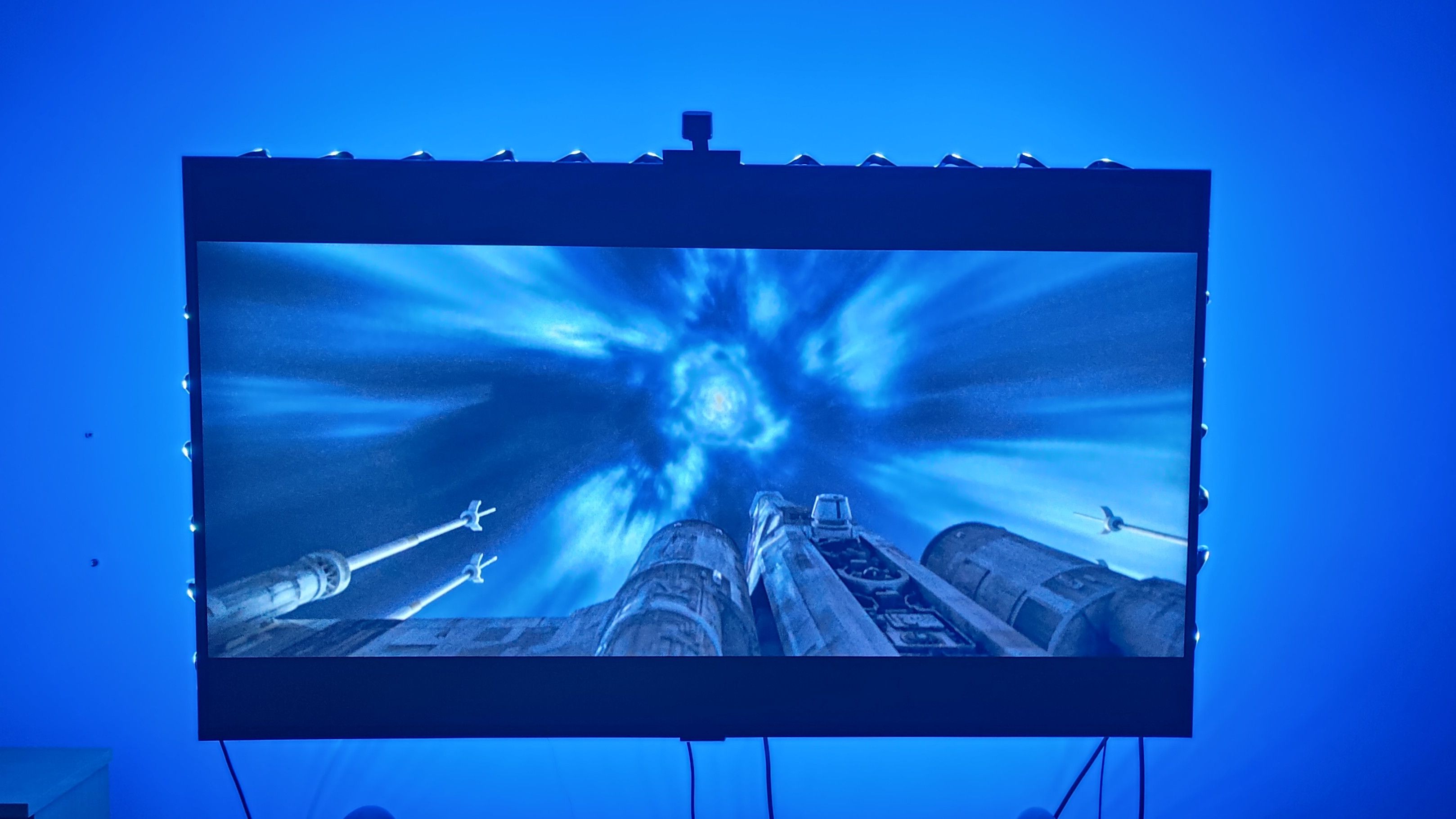 An X-Wing beams into hyperspace in Rogue One, while the TV&#039;s LED lightstrips project a blue hue onto the wall behind.