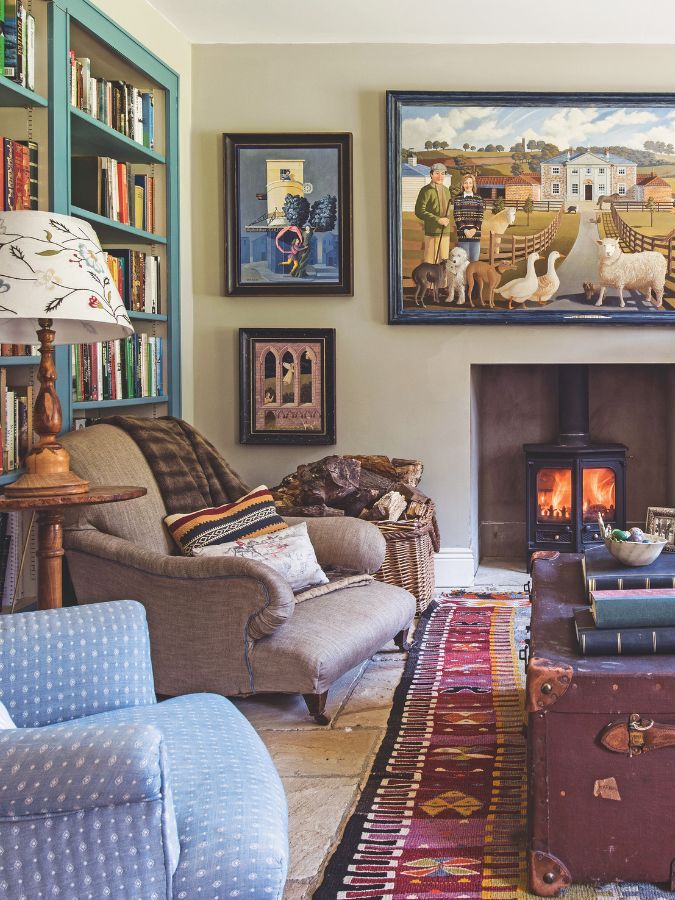 A cozy and eclectic living room with a library wall, lit wood burner, patterned large rug, vintage look trunk and a mismatched upholstered sofa and chairs.