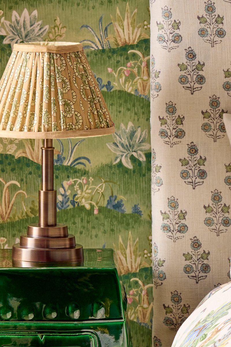 Beige and green patterned table lamp pictured in a green wallpapered bedroom that features a bed with a botanically patterned headboard and bedding.