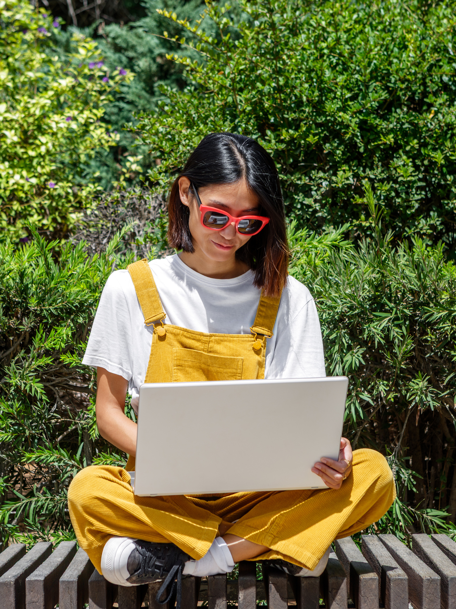 A woman sits cross-legged on a bench, wearing yellow overalls and sunglasses, while focused on her laptop. The setting is a lush green garden, indicating a warm, sunny day.