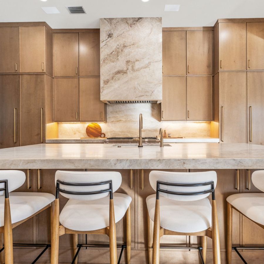 Natural wood cabinetry in a wood and warm marble kitchen, with floor to ceiling cabinetry and large island with four white upholstered bar stools. There is a potted tree visible in the background