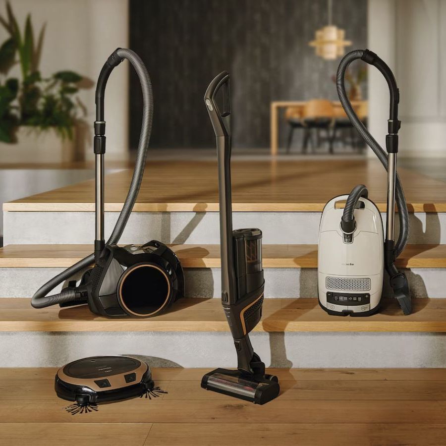 Four Miele vacuum cleaners &amp;ndash; a cordless vacuum, robot vacuum and two canister vacuums &amp;ndash; on set of wooden stairs in luxury home, with plants and furniture pictured in background.