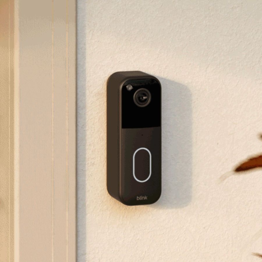 Blink Video Doorbell on cream wall next to beige door with black lock, with blurry leaves pictured to the right.