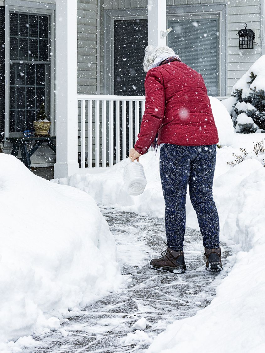 A senior adult woman is sprinkling/spreading de-icing salt crystals on her home's front walkway to melt the slippery ice and snow.