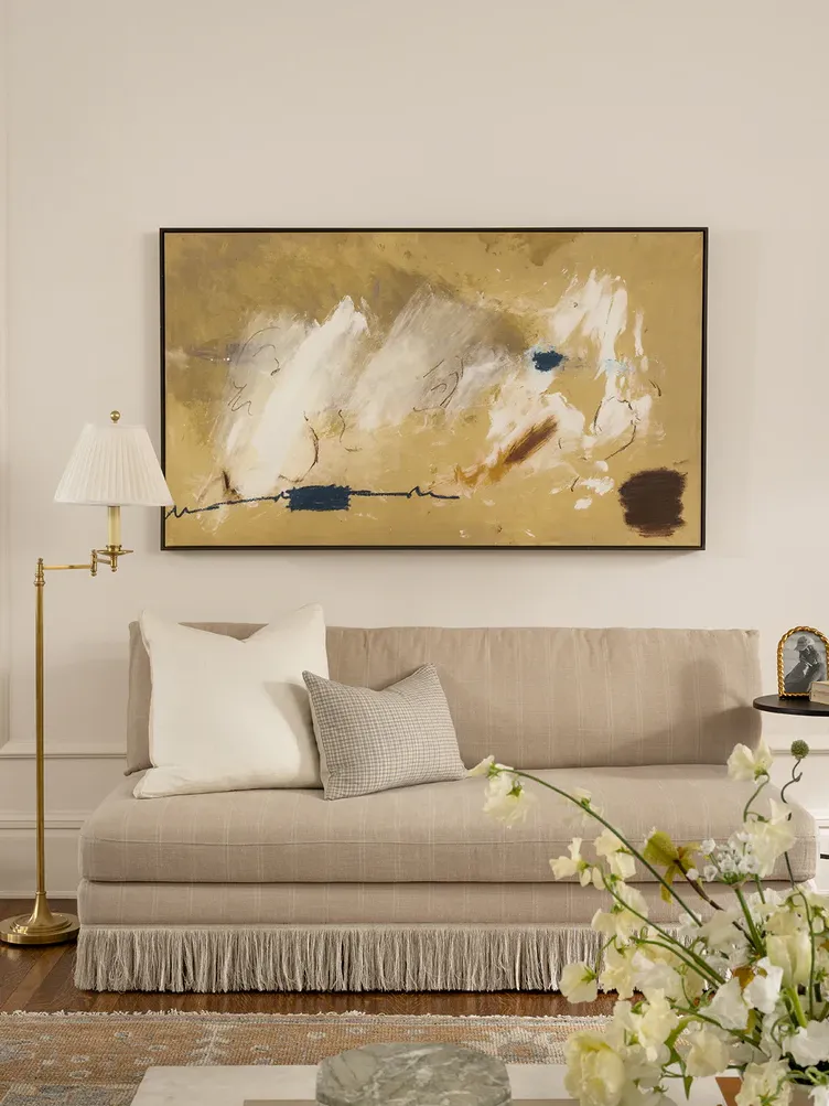 A neutral living room with a neutral armless couch with a fringed base