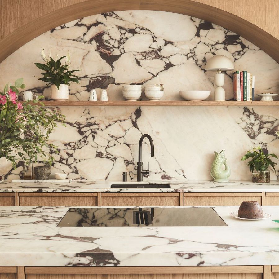 A modern kitchen with arched lightwood fittings over a kitchen sink, with a large marble backsplash. An island opposite with matching marble top, and a glass stove top. A vase of roses to the left.