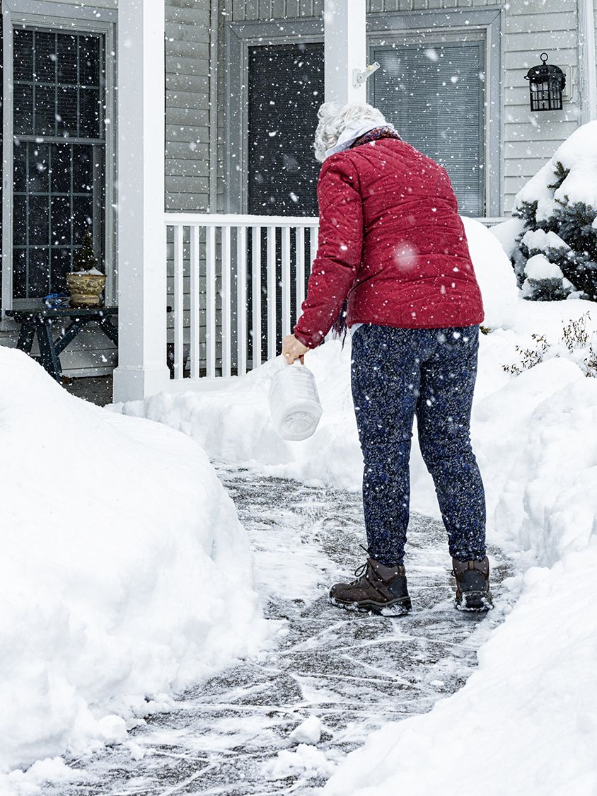 A senior adult woman is sprinkling/spreading de-icing salt crystals on her home's front walkway to melt the slippery ice and snow.