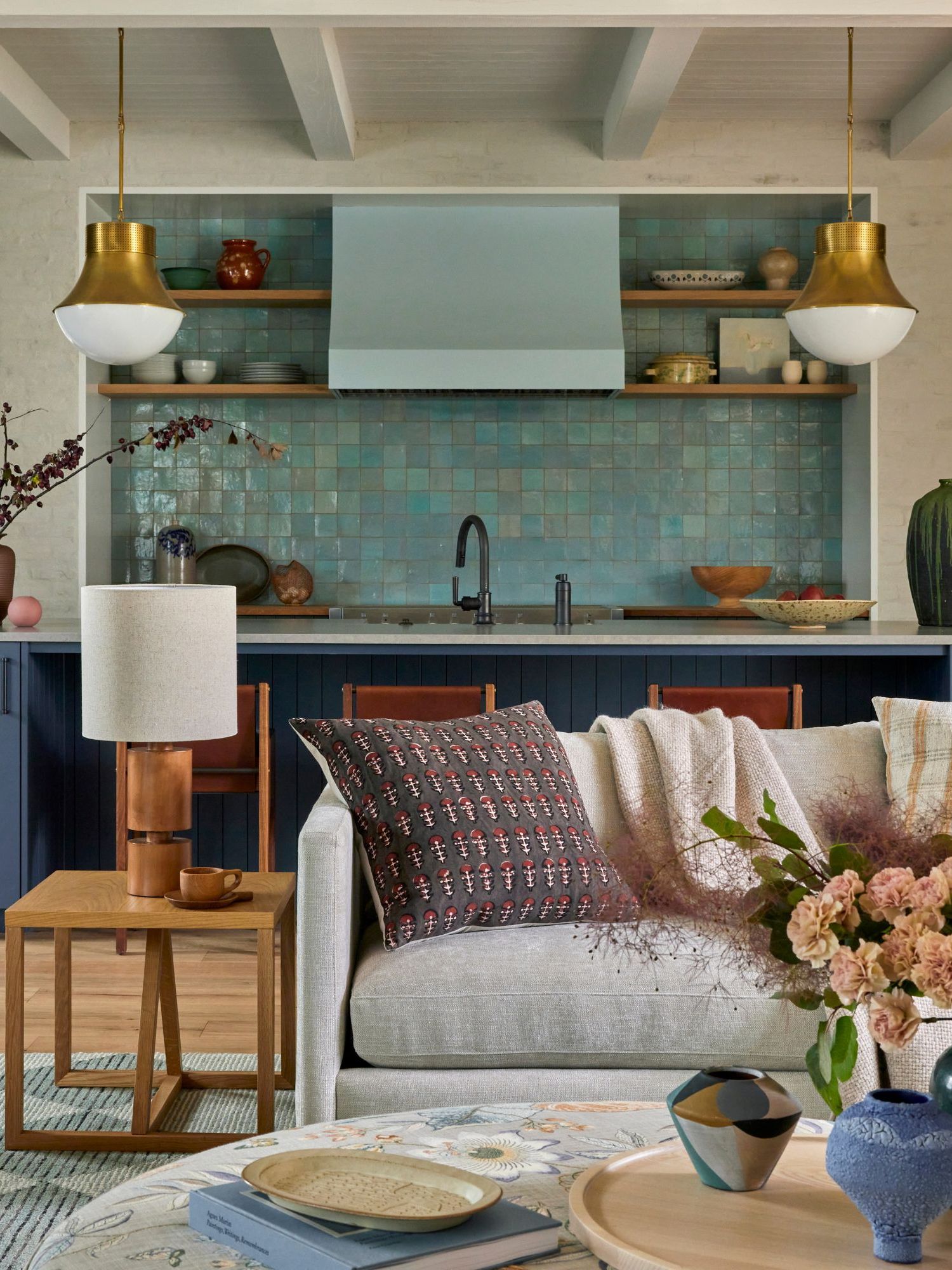 A modern, open-plan living room and kitchen. A teal blue tiled backsplash, dark blue cabinets, rustic decor, and two brass pendant lights. A gray sectional with patterned scatter cushions, a round coffee table with a tray and a vase of flowers, and a wooden side table with a table lamp.