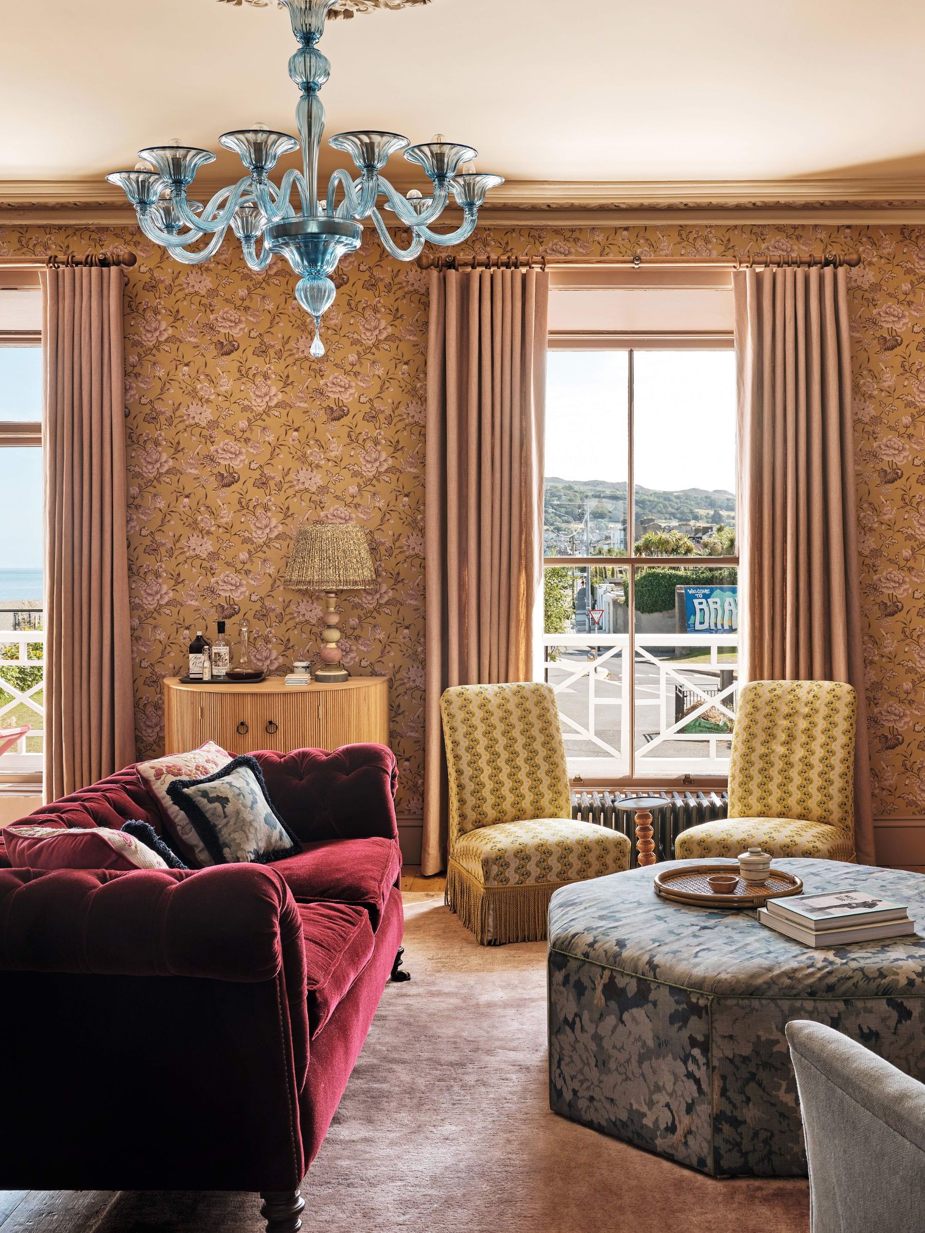 a sunlit sitting room with peach toned floral wallpaper, burgundy velvet couch, yellow slipper chairs, tapestry ottoman and a blue murano chandelier