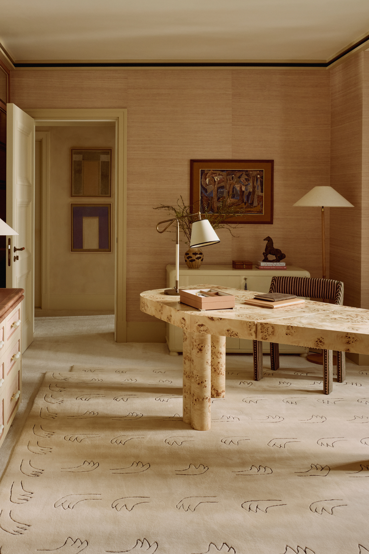 Neutral office space featuring an asymmetrical burl wood desk, linen-shaded table lamps, wall art, textured wallpaper, a figural motif beige rug, and a striped office chair