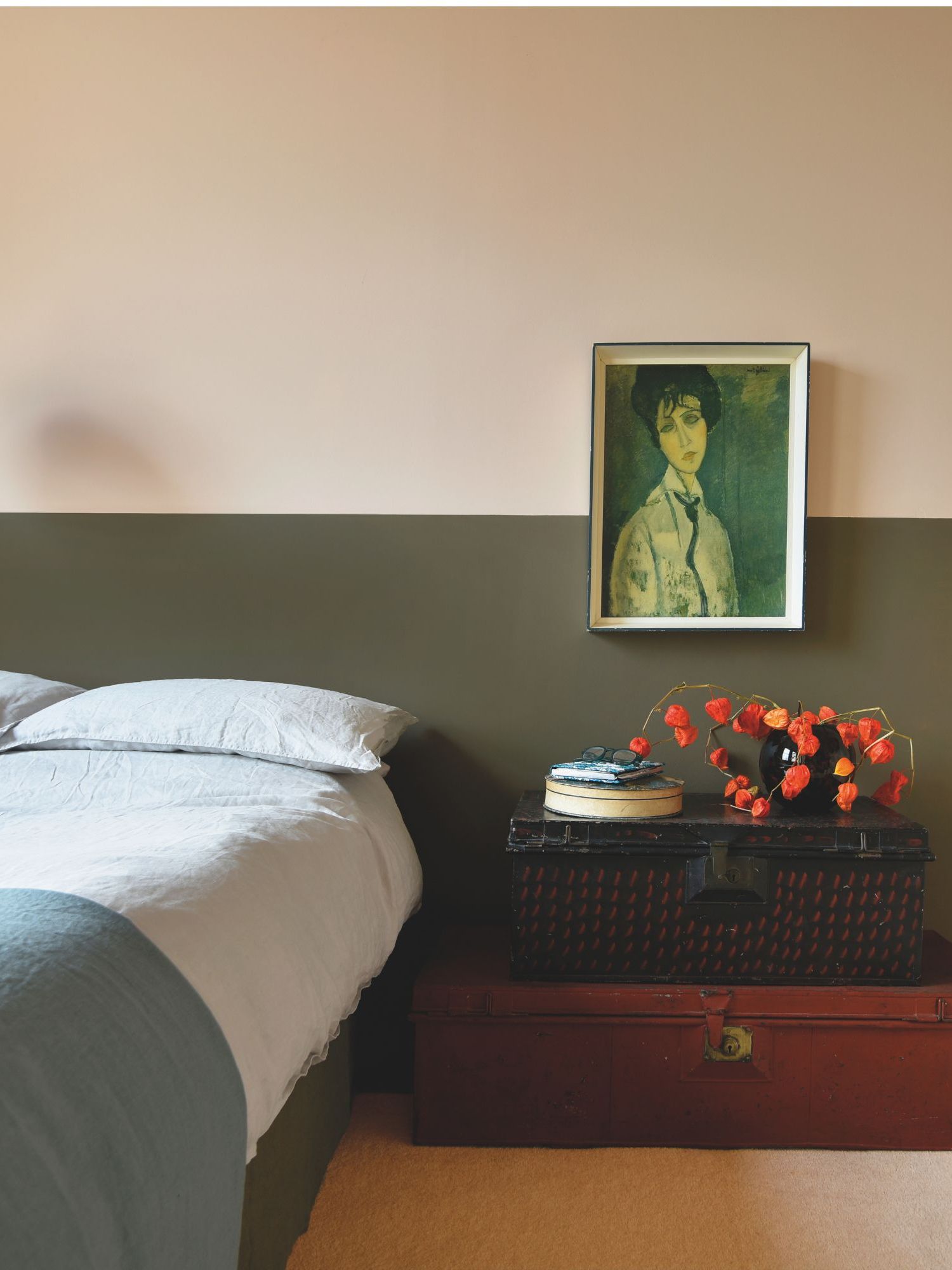 Bedroom with wall painted half pale pink and half dark brown, a low bed with pale blue linen bedding, two vintage trunks act as a nightstand with a vintage print placed above and a vase of chinese lanterns