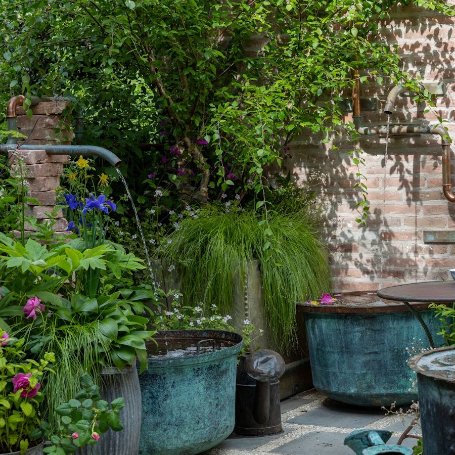 Courtyard garden with water pipes flowing into large blue copper pots and lush, cottage-style planting