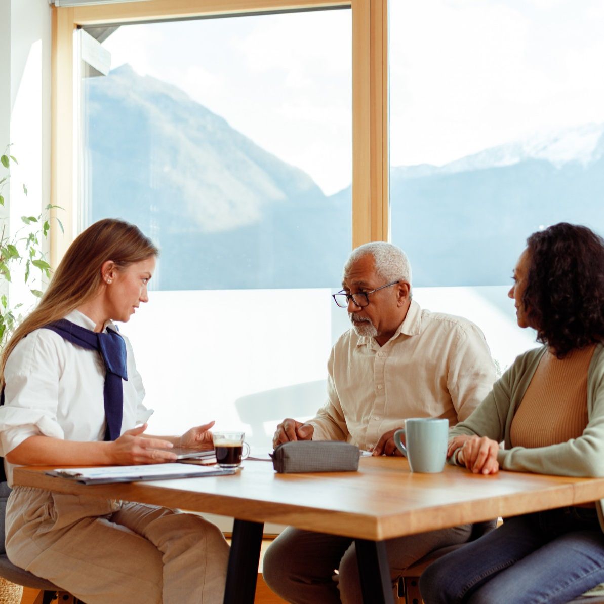 A couple sits at their kitchen table with a dramatic view of mountains. They are meeting with a younger woman with paperwork.