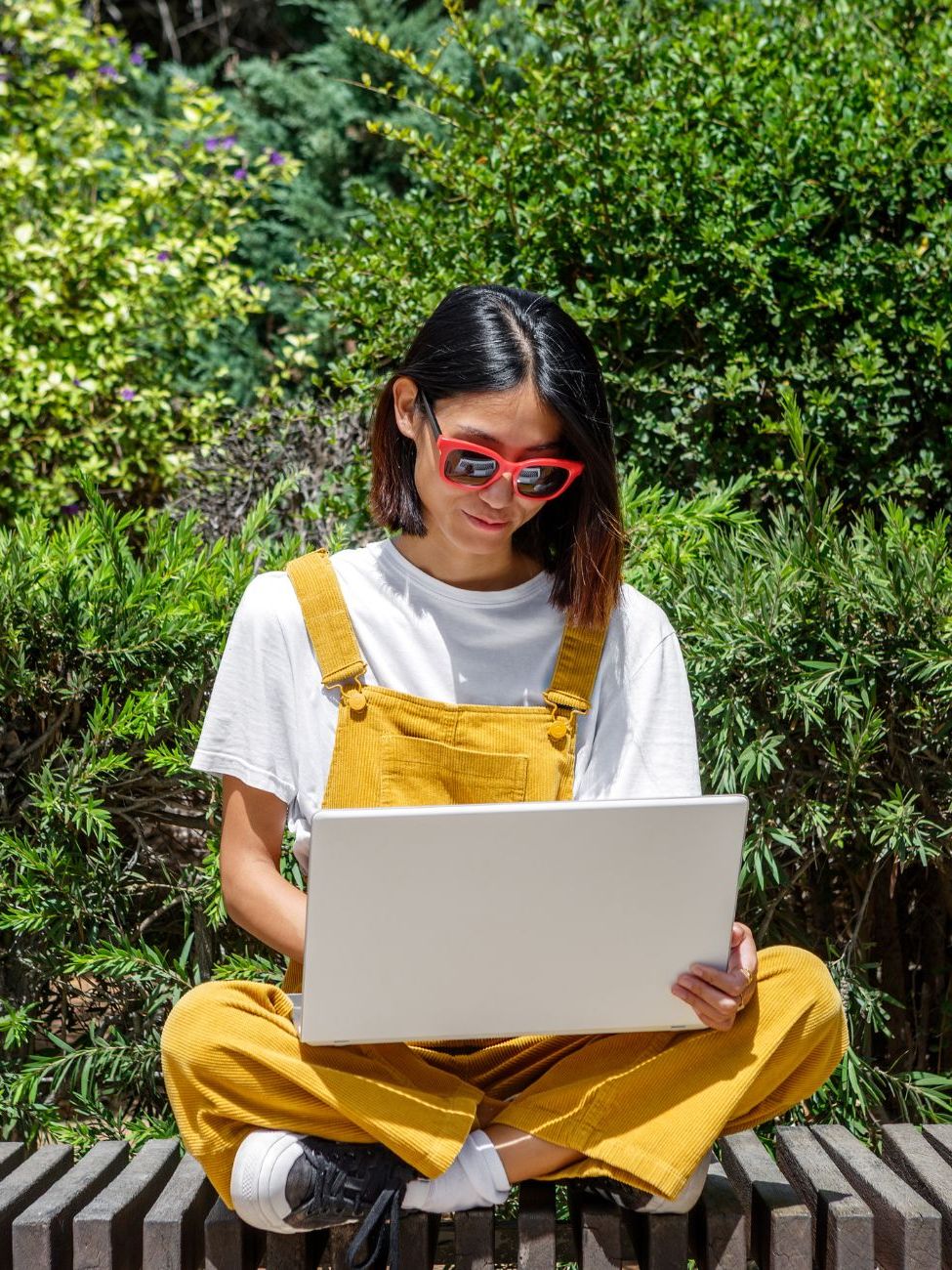 A woman sits cross-legged on a bench, wearing yellow overalls and sunglasses, while focused on her laptop. The setting is a lush green garden, indicating a warm, sunny day.
