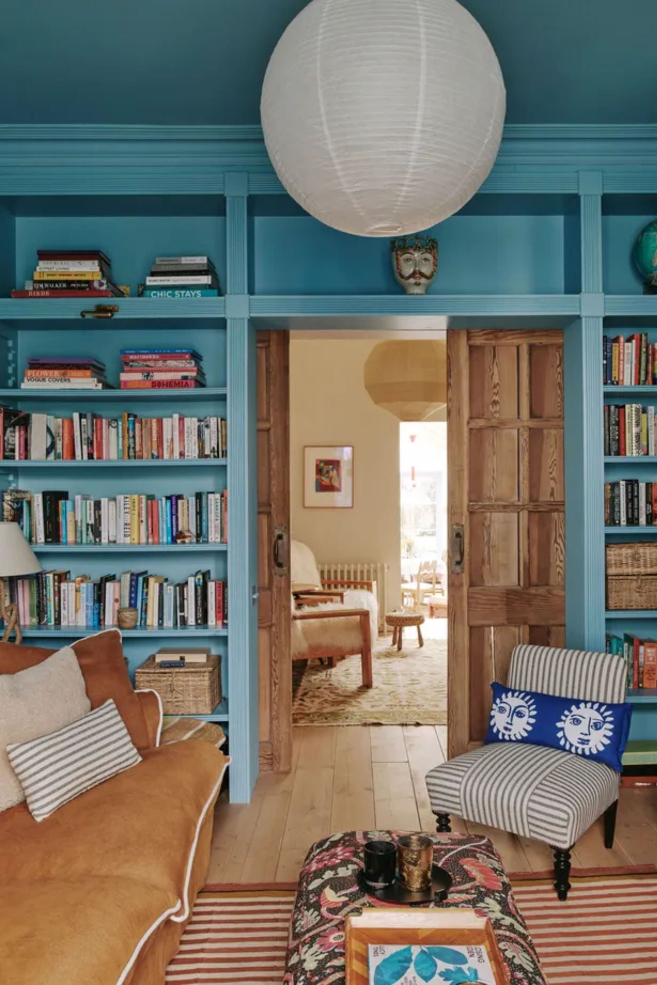 A cozy living room with blue color-drenched walls with built-in shelving, a brown sofa, a striped rug, and a patterned ottoman.