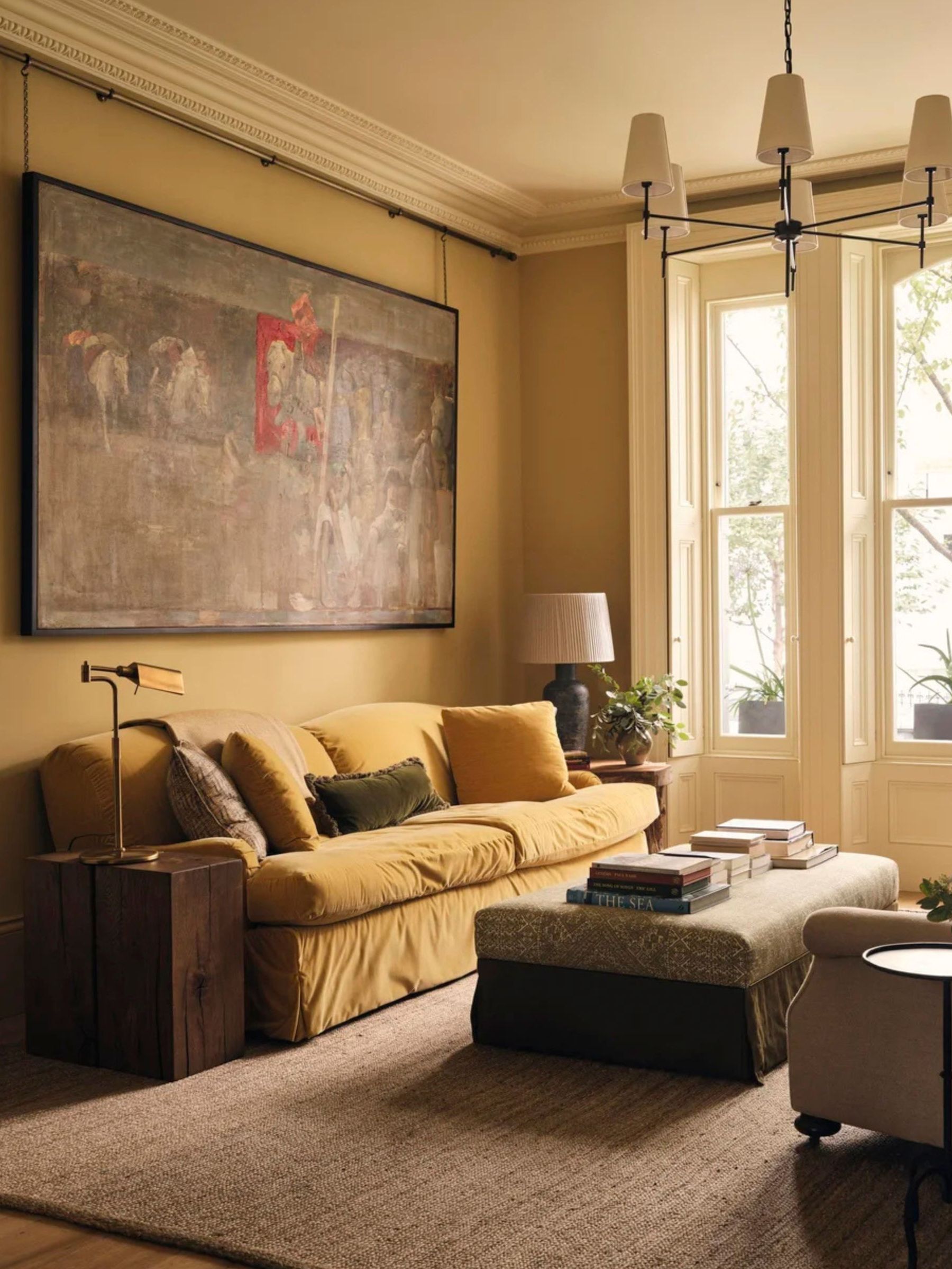 A warm-toned living room with ochre walls features a yellow velvet sofa, a large vintage painting hanging on a rail, and an ottoman coffee table in front of tall bay windows