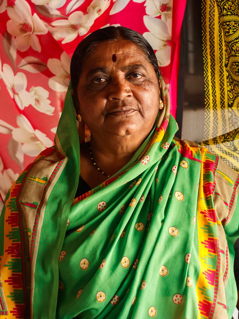 A woman wearing a green and red patterned sari looks at the camera, standing in front of a wall decorated with two red and gold saris.