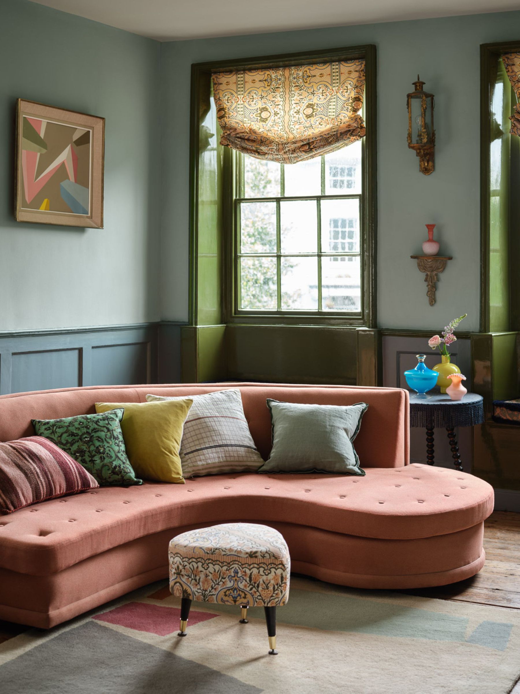 Blue painted living room with curved sofa and vintage accents including heart shaped foot stool and patterned window treatments.