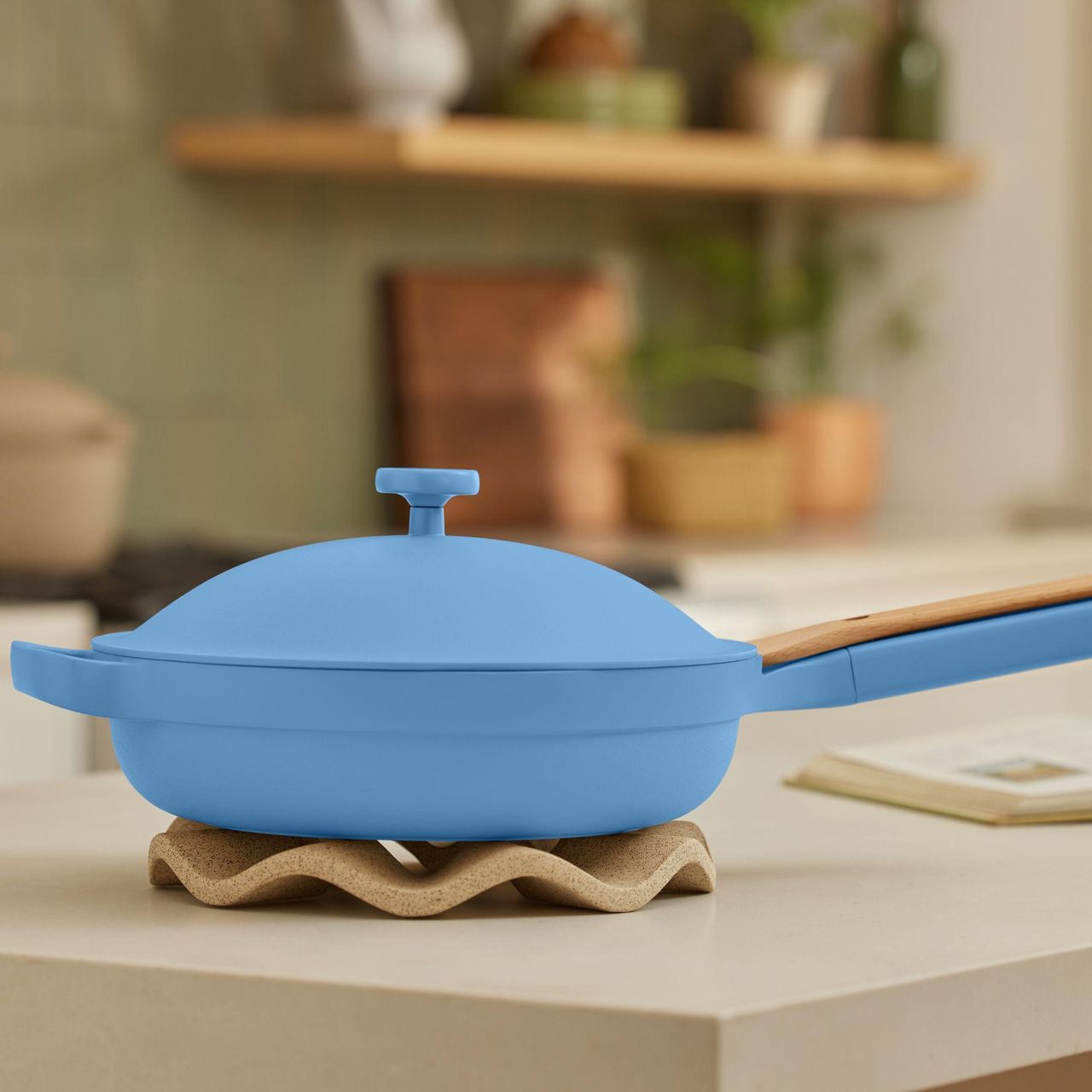 A blue pan with a long handle on one side and a carry handle opposite. A matching domed lid sits on top. The pan sits on a wavy trivet on a white quartz countertop, and there is an open cookbook out of focus to it's right.
