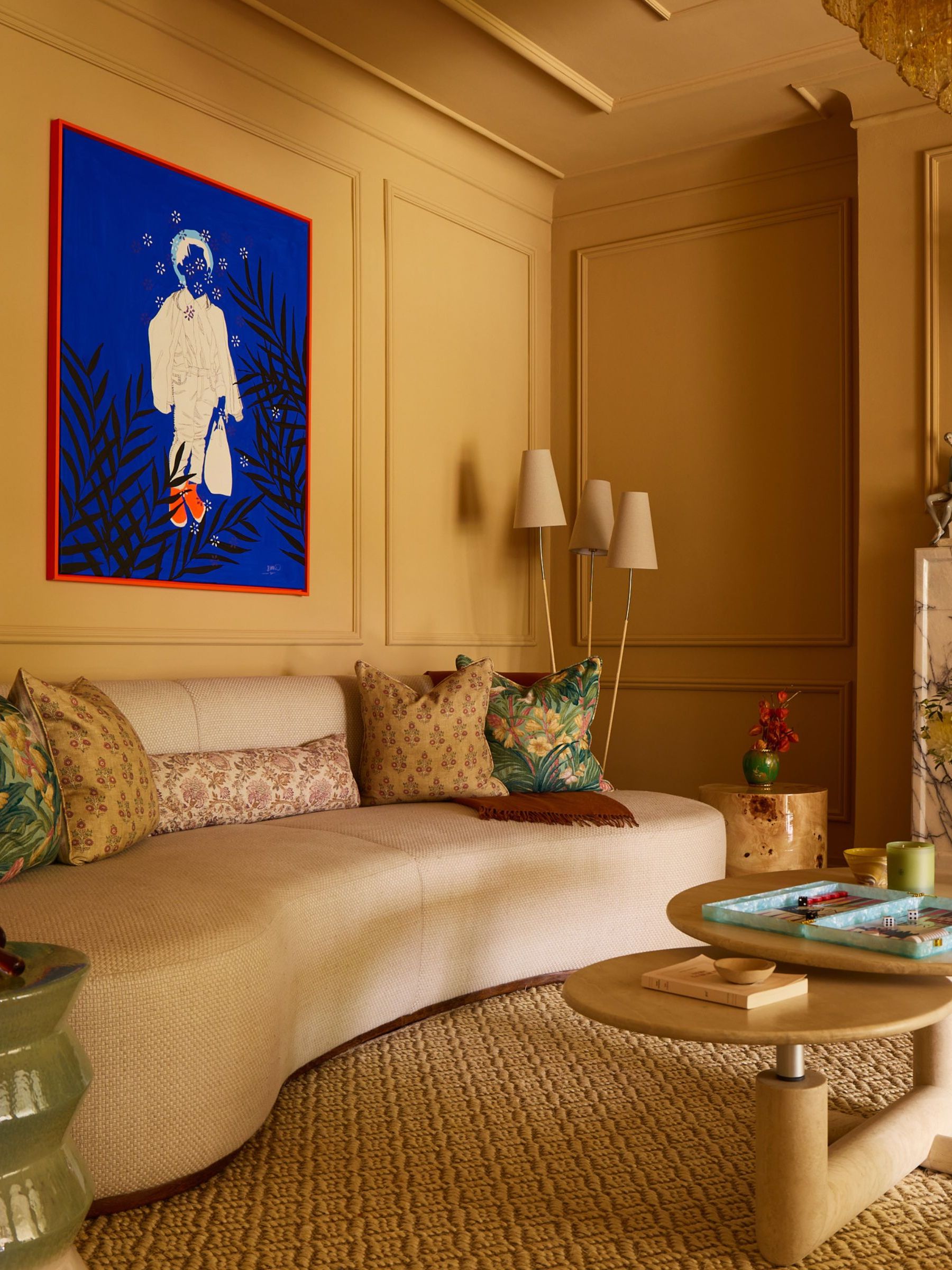 Living room with deep mustard walls, marble fireplace, a large cream curved sofa decorated with block-printed cushions, a round tiered coffee table, and a large cobalt blue painting on the wall.